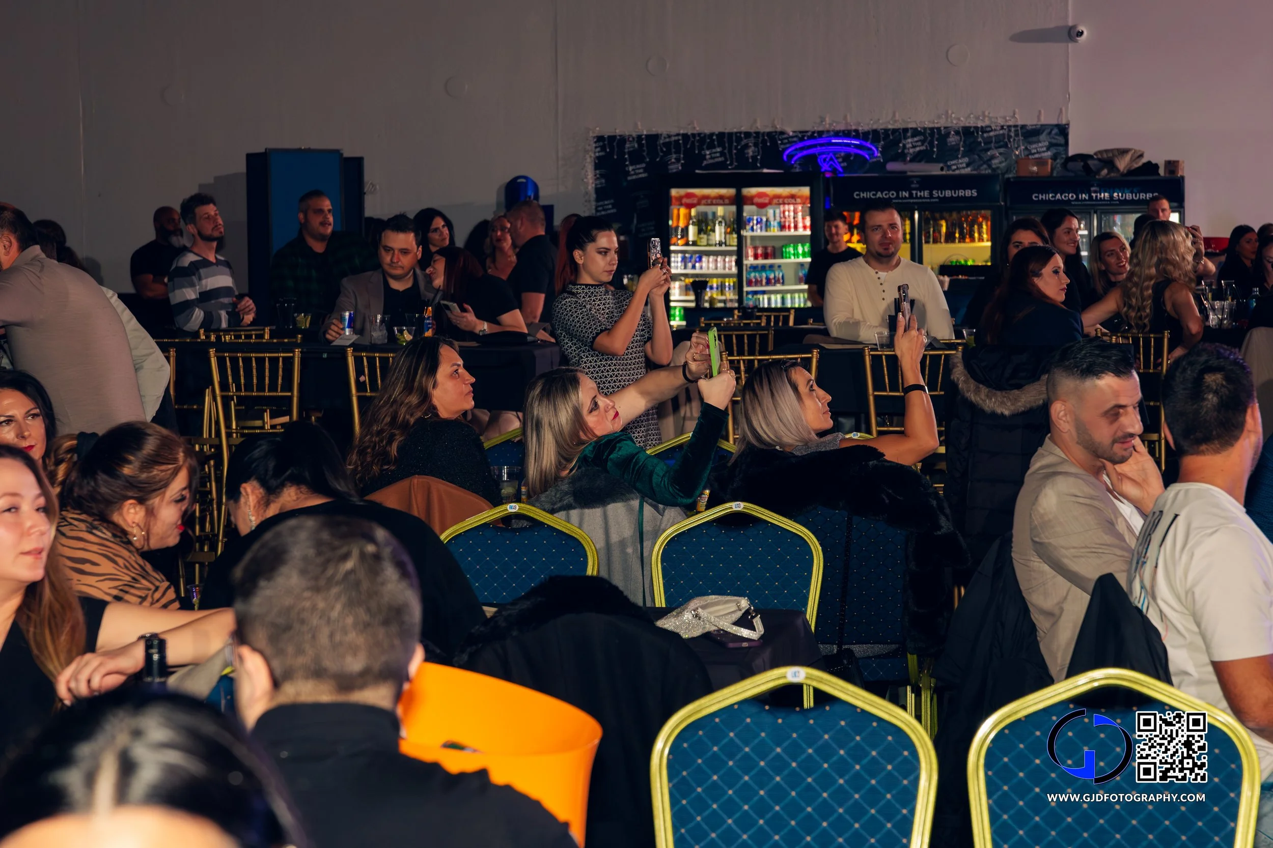 People sitting and standing at tables in a conference or event space, some taking photos or videos, with a bar in the background and a dark ceiling.