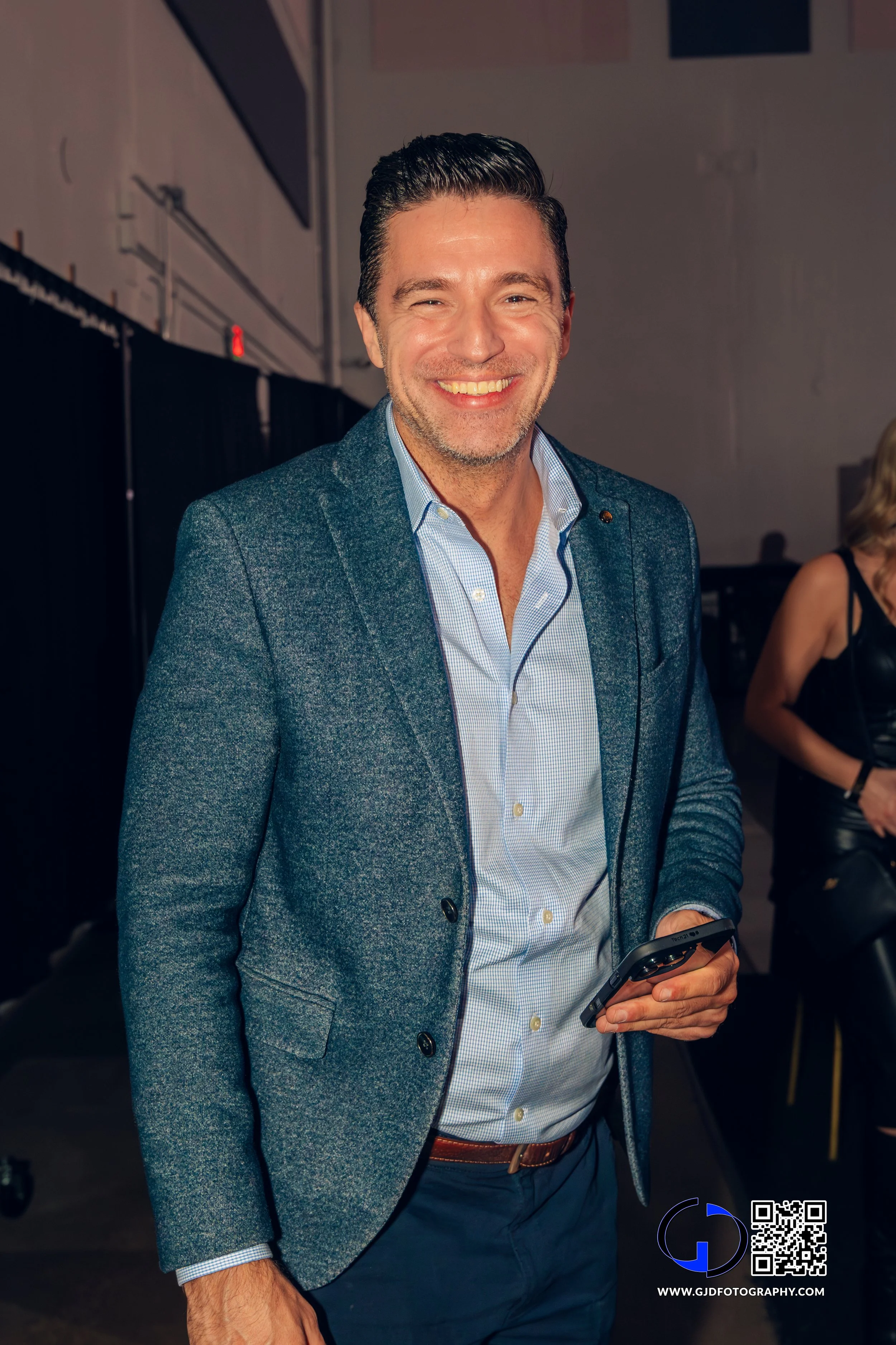 Smiling man in a blue blazer and dress shirt holding a smartphone at an indoor event.