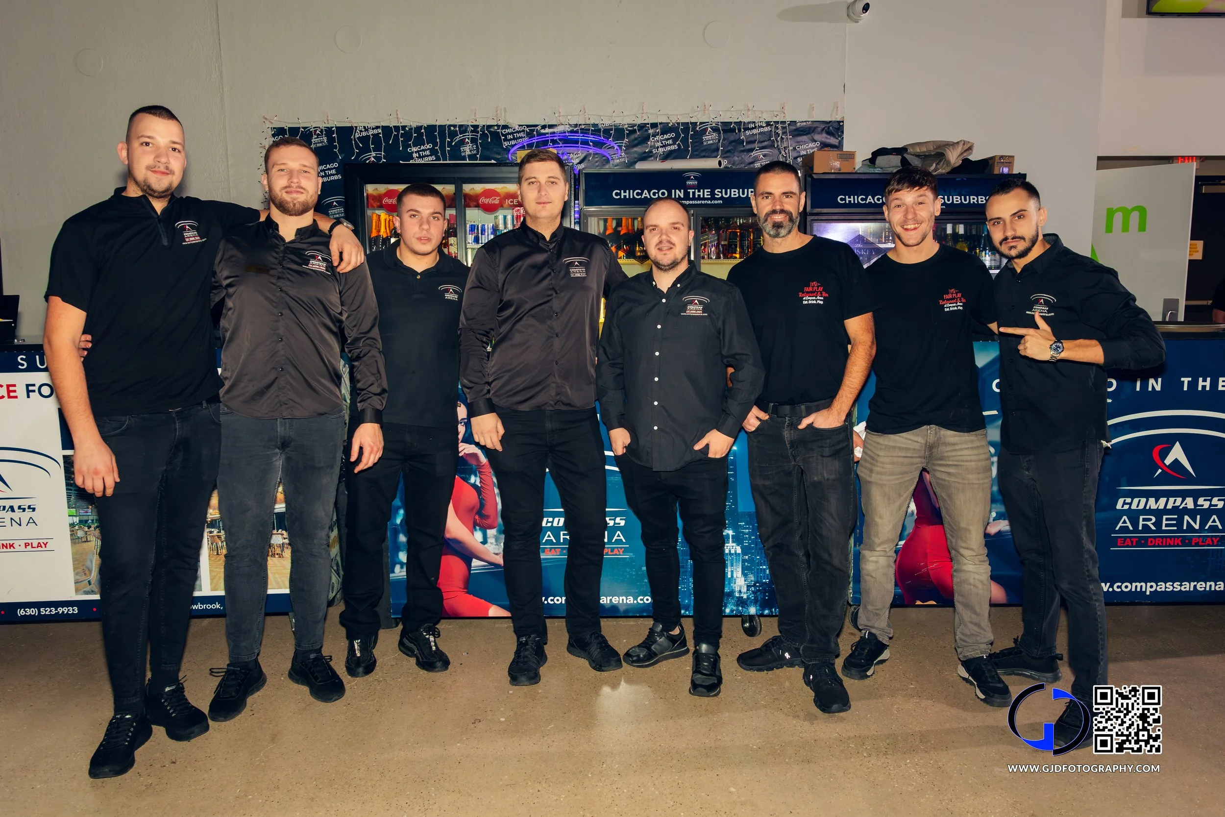 Group of eight men standing together in front of a vending area at Compass Arena, wearing black shirts and pants, posing for a photo indoors.