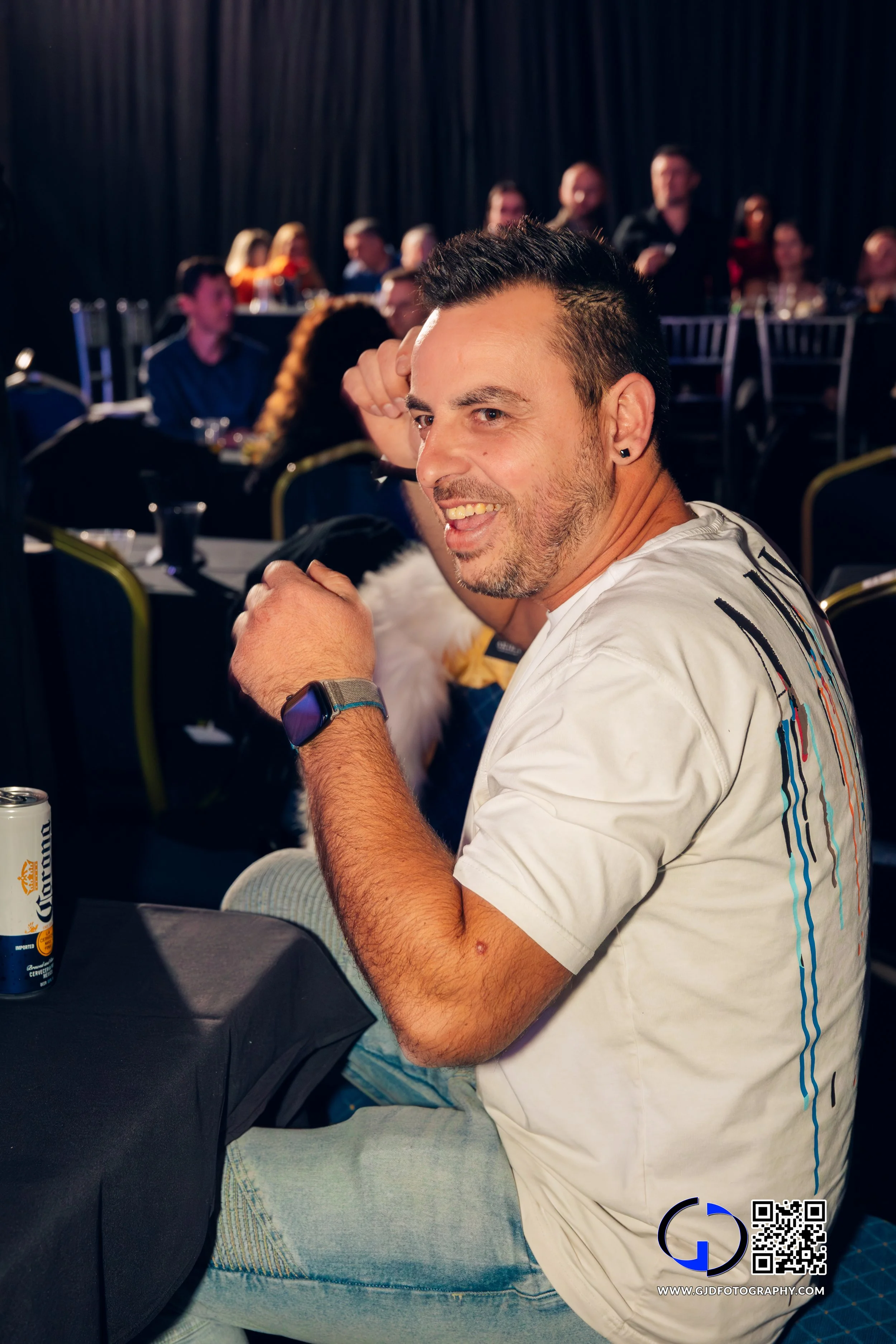 Man smiling and sitting at a table during a formal event or celebration, with other people seated in the background.