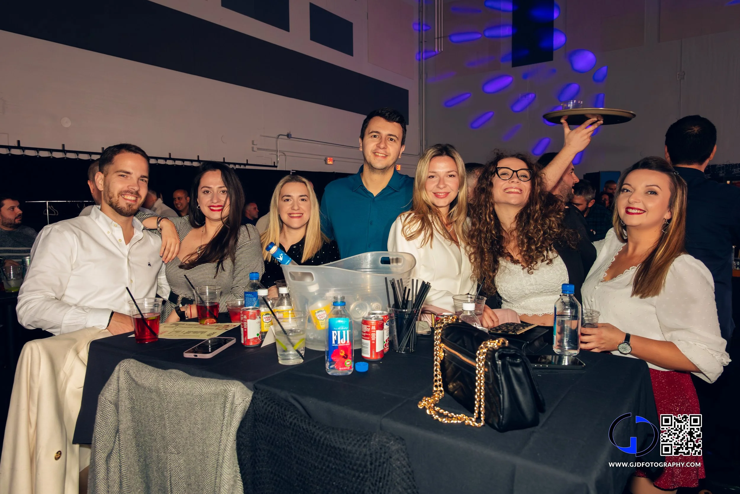 Group of seven friends smiling and posing behind a table at a party or event, with drinks, snacks, and a black purse on the table, in a venue with purple lights and a large crowd in the background.