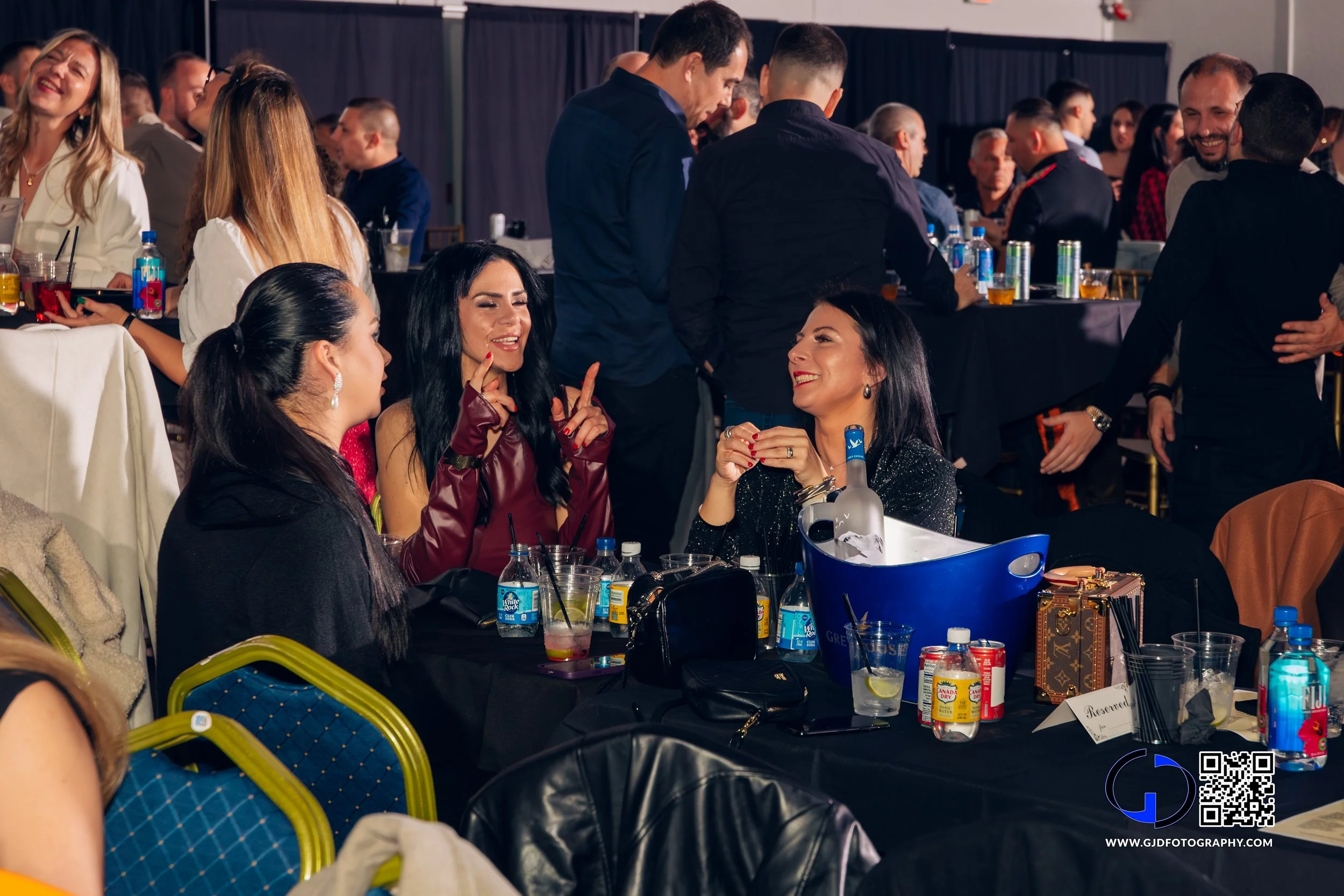 People socializing at a formal event or party, seated around tables with drinks and snacks, with some standing and chatting in the background.