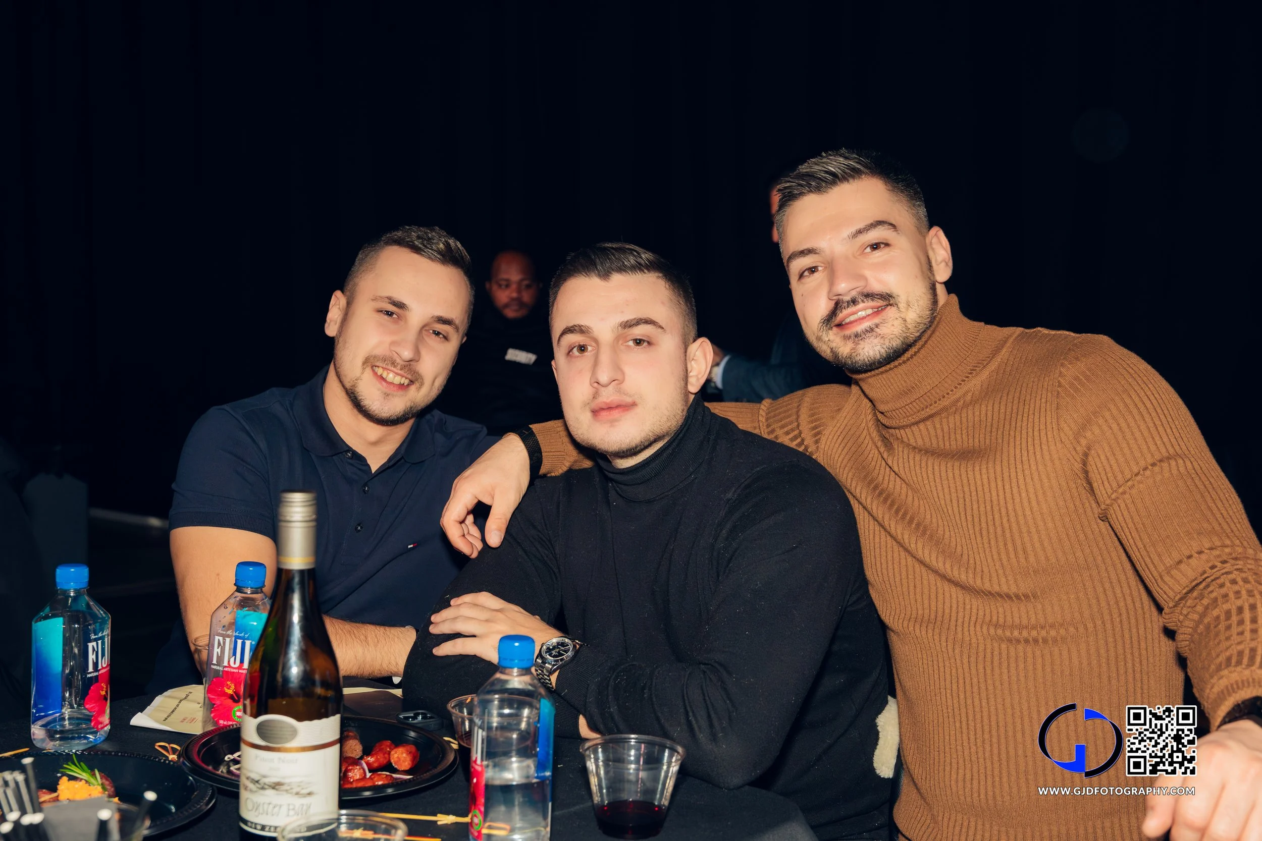 Three men sitting at a table, smiling at the camera, with food and drinks on the table. Two are wearing dark shirts, one in a brown turtleneck, in a dimly lit setting.