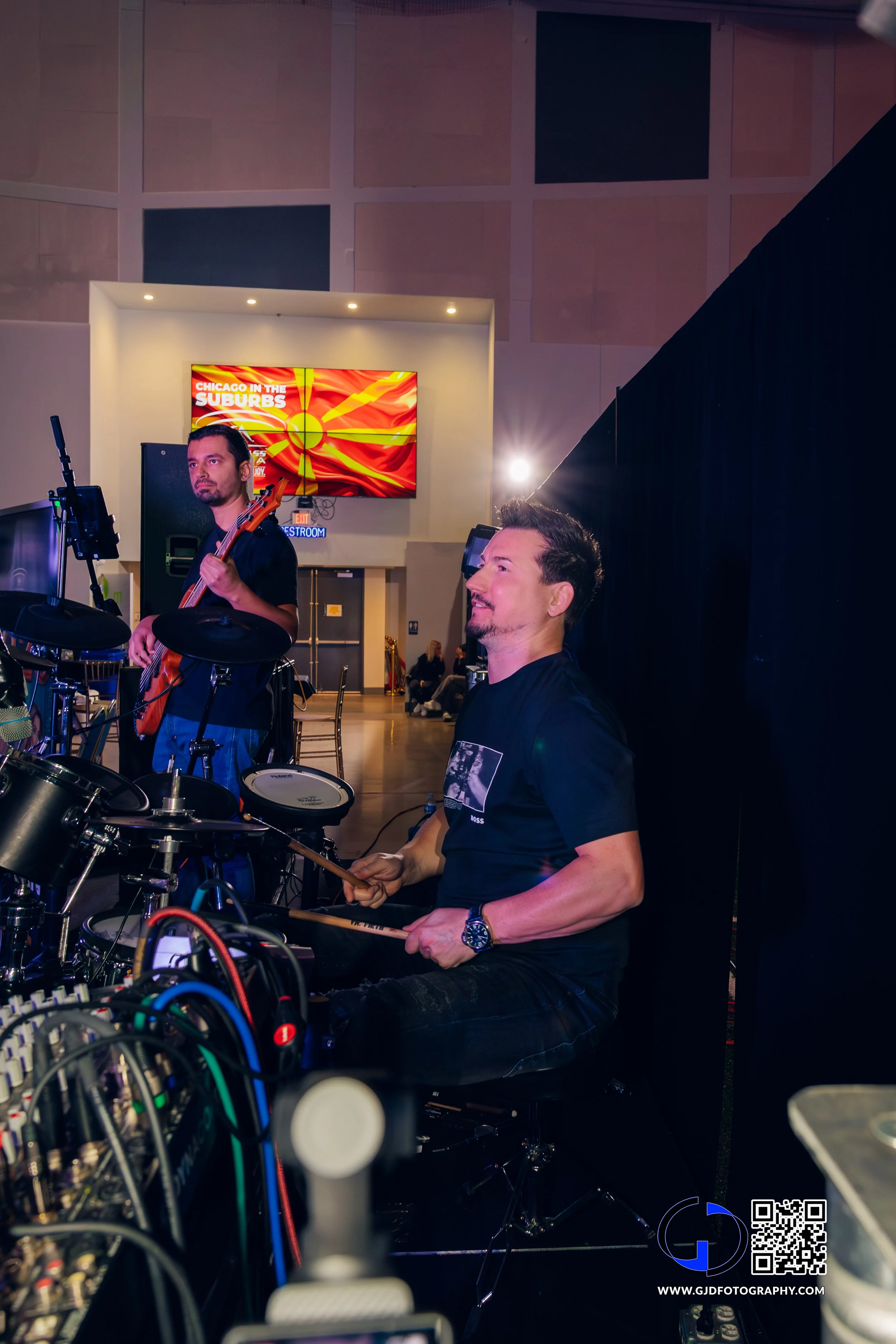 Two musicians with instruments and audio equipment backstage at an event, one playing drums and the other holding a bass guitar, with a digital display in the background.