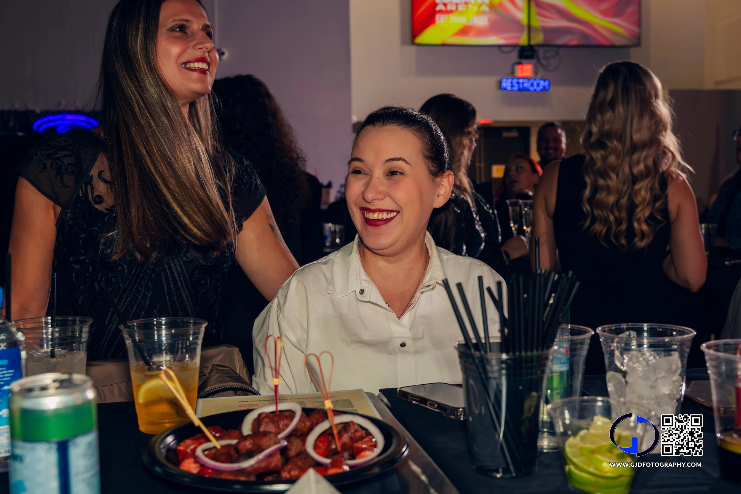Two women smiling and chatting at a table during a celebration or party, with food, drinks, and party supplies in front of them.