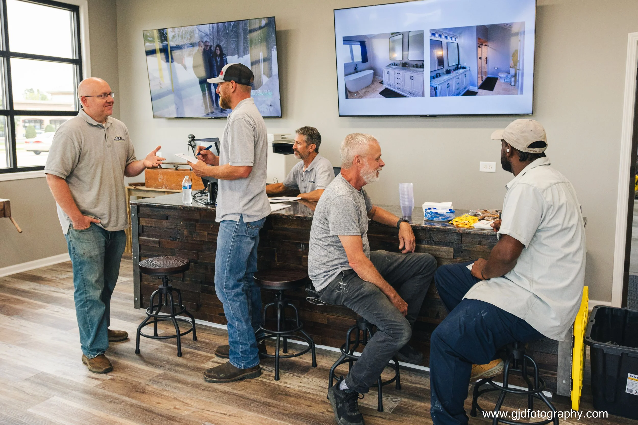 Five men are inside a modern office or showroom, engaged in conversation. Two are standing by a dark wooden counter, one is seated on a stool, and two others are seated on the counter. There are large screens on the wall displaying interior design im