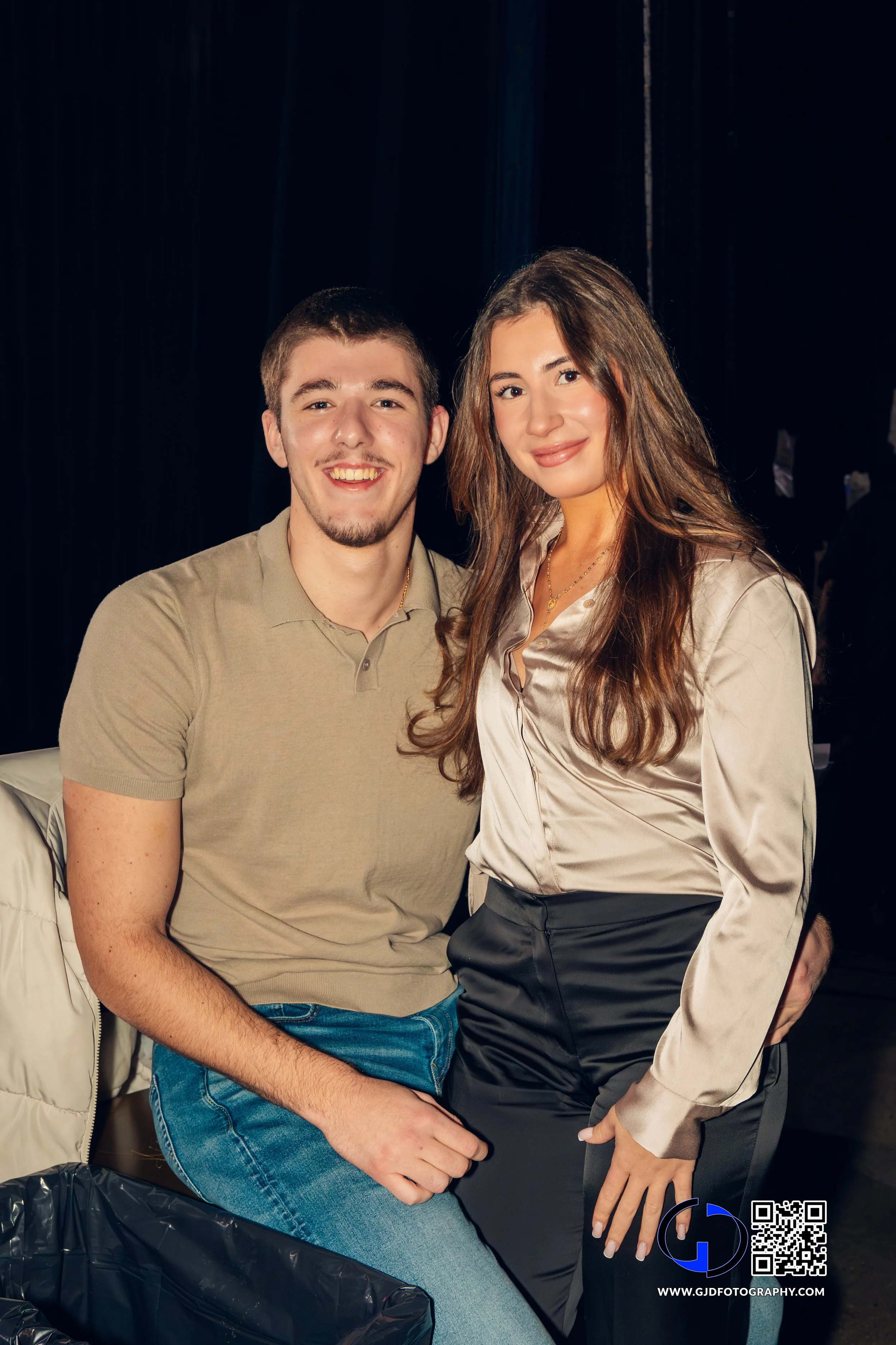 A young man and woman smiling and posing together, with the man sitting and the woman standing, in an indoor setting with black background curtains.