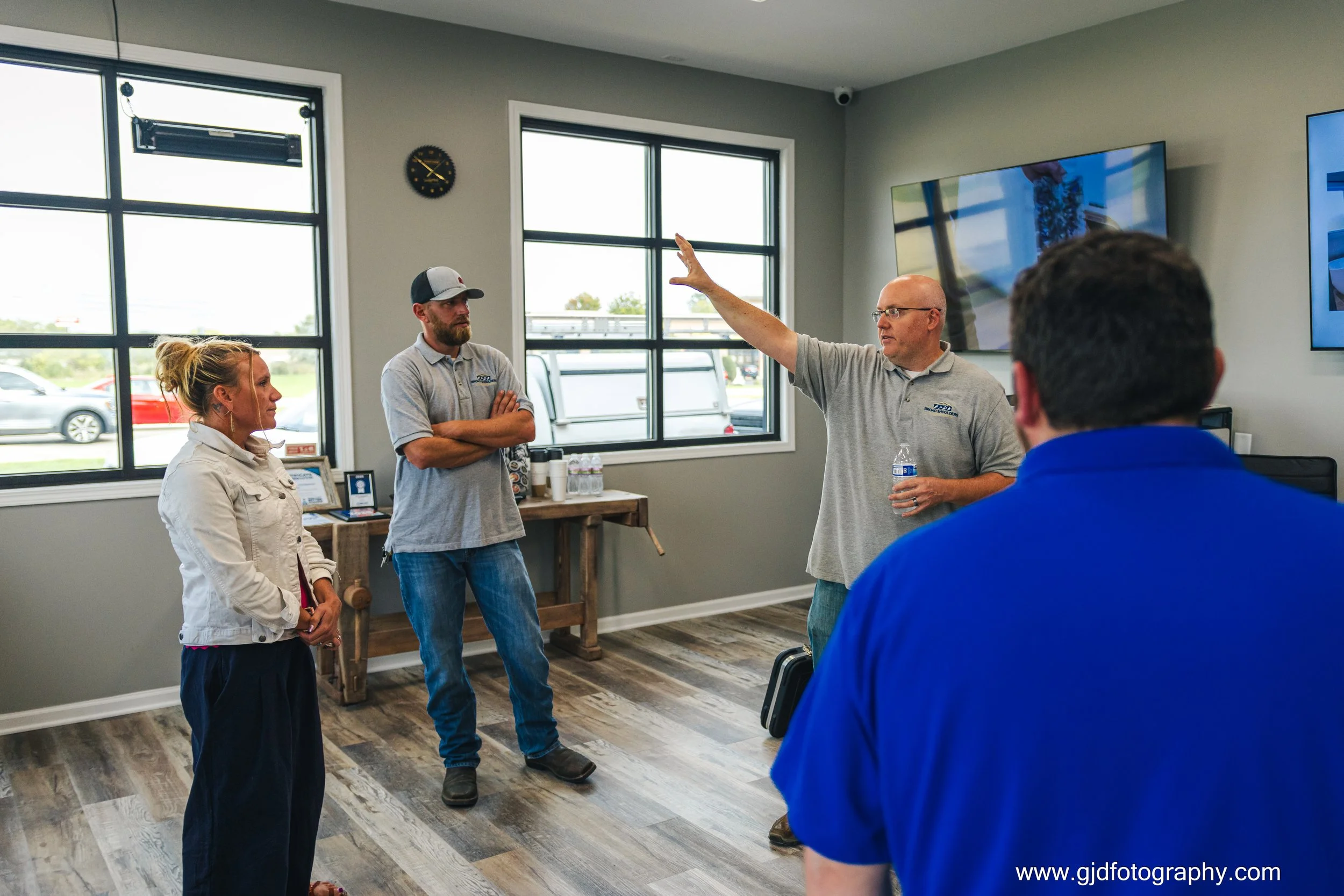 A man is giving a presentation to a group of four people in a modern office with large windows and wood flooring. The presenter is bald, wearing glasses and a gray polo, holding a water bottle, and gesturing with his right hand. The group includes a 