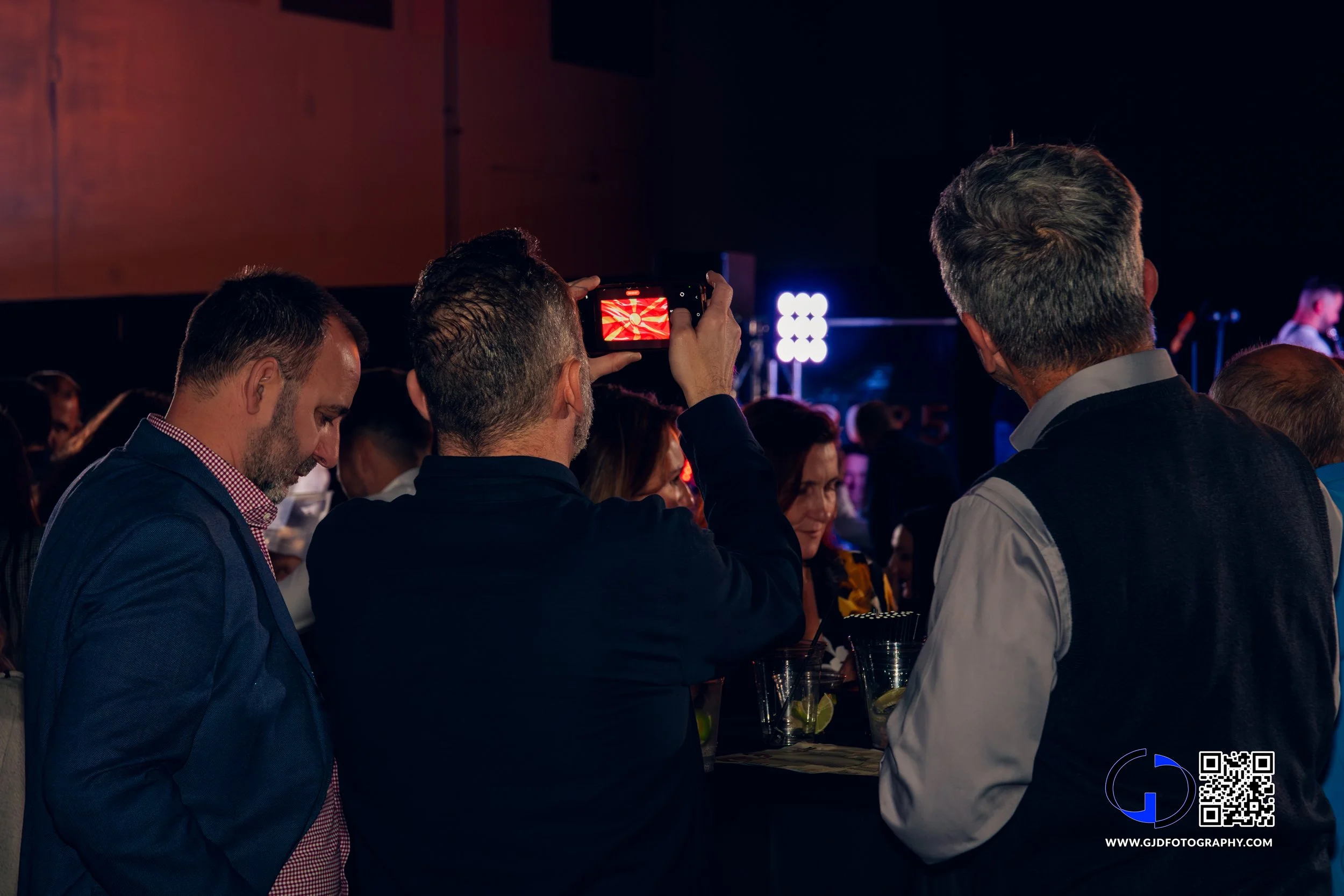 Group of people at a party or event, with one person taking a photo on a smartphone, in a dimly lit setting with stage lights and drinks on the table.