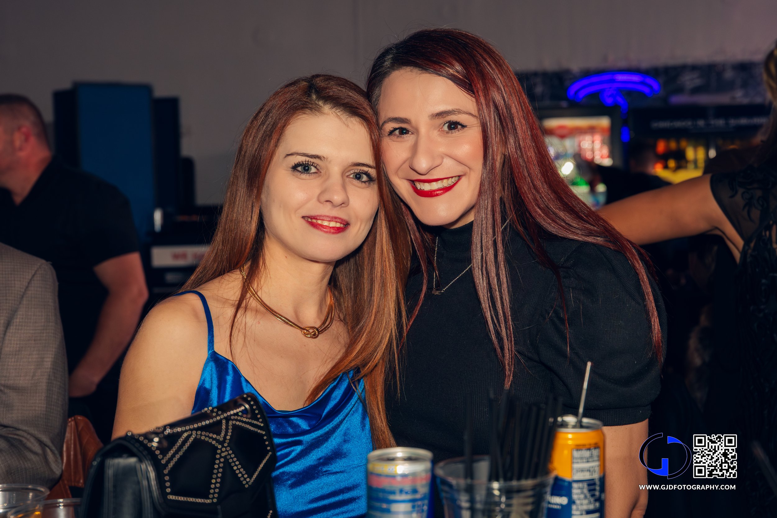Two women with red hair smiling at a social event, one in a blue dress and the other in a black outfit, sitting at a table with drinks.