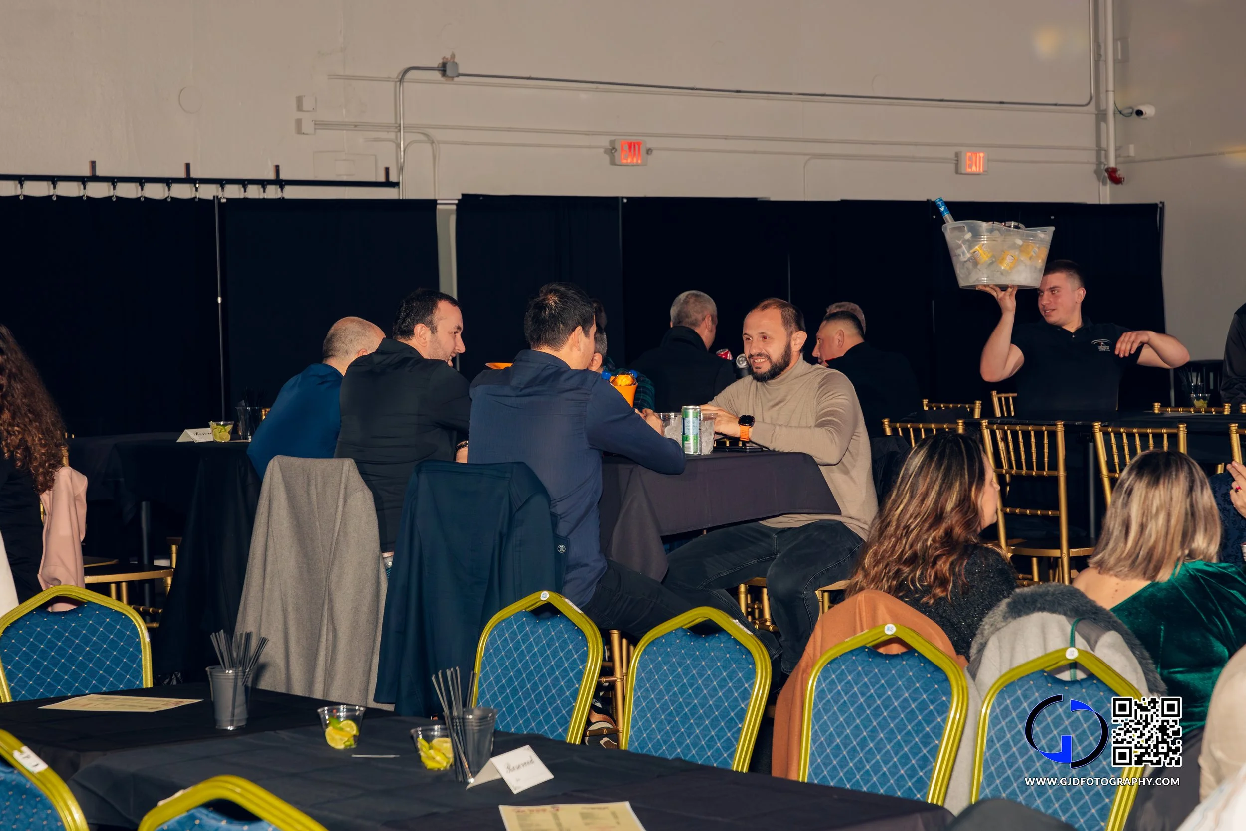 People sitting at a table in a banquet hall, engaged in conversation, with a waiter holding a large drink bucket in the background.