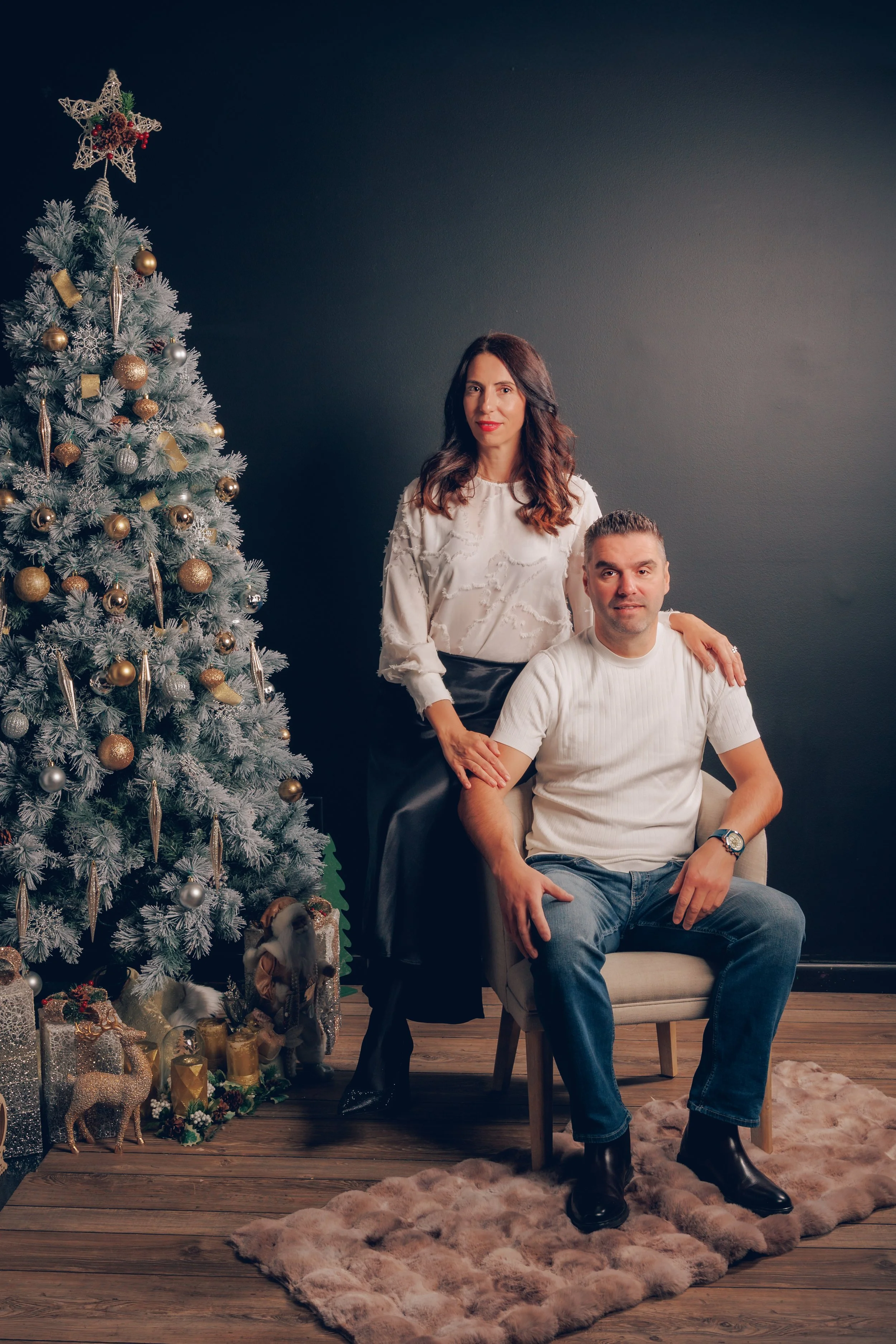 A woman with brown hair and a white blouse standing behind a seated man with short hair wearing a white shirt, in a room decorated for Christmas with a Christmas tree, ornaments, and presents.