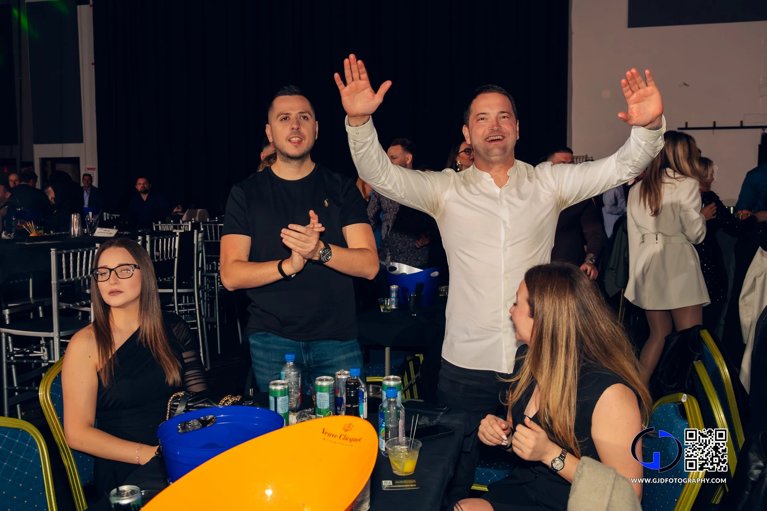 People at a party, some standing and clapping, others sitting, with drinks on the table, in a dimly lit room.