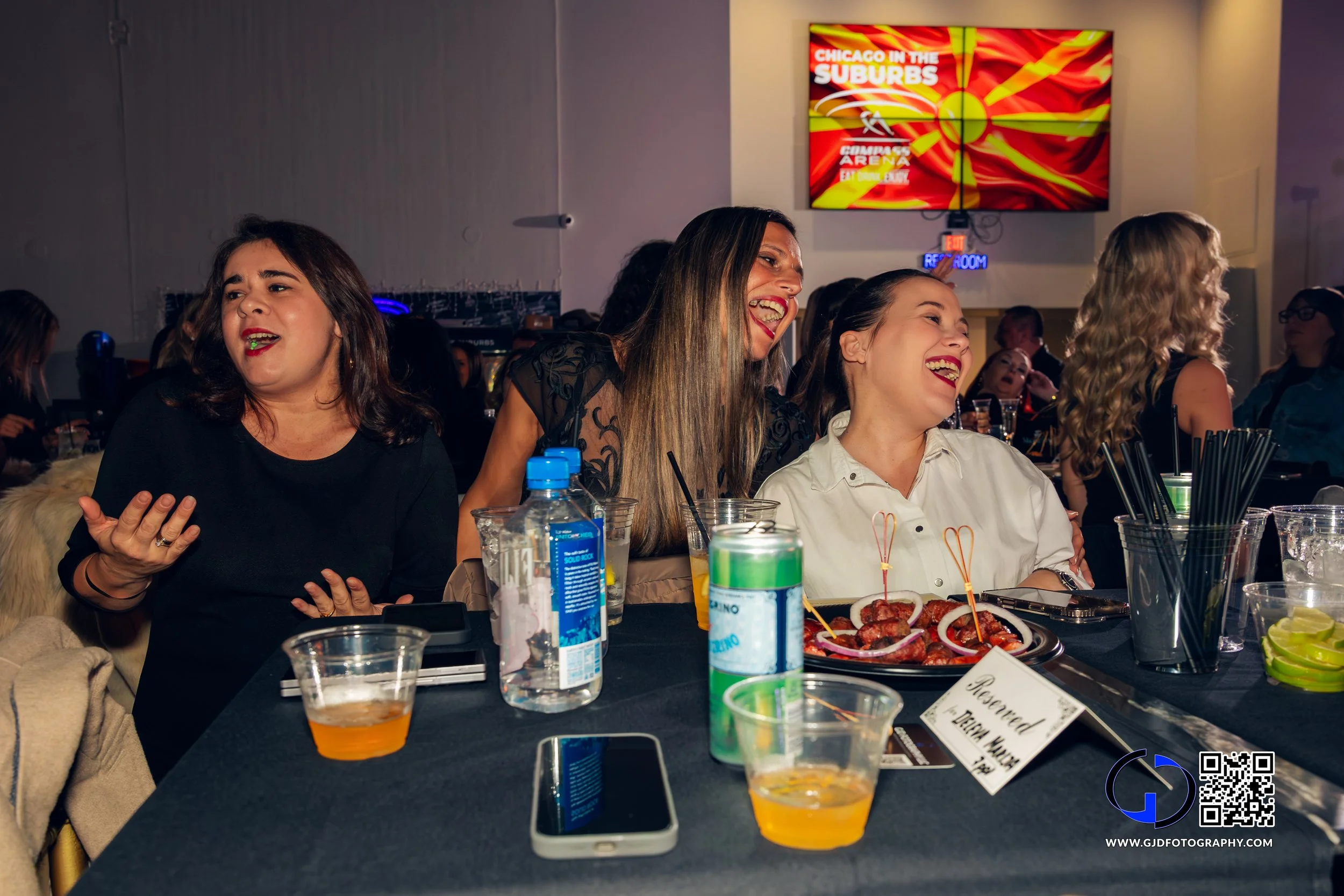 Three women sitting at a bar or restaurant, laughing and enjoying drinks and food, with a TV screen in the background displaying colorful graphics.