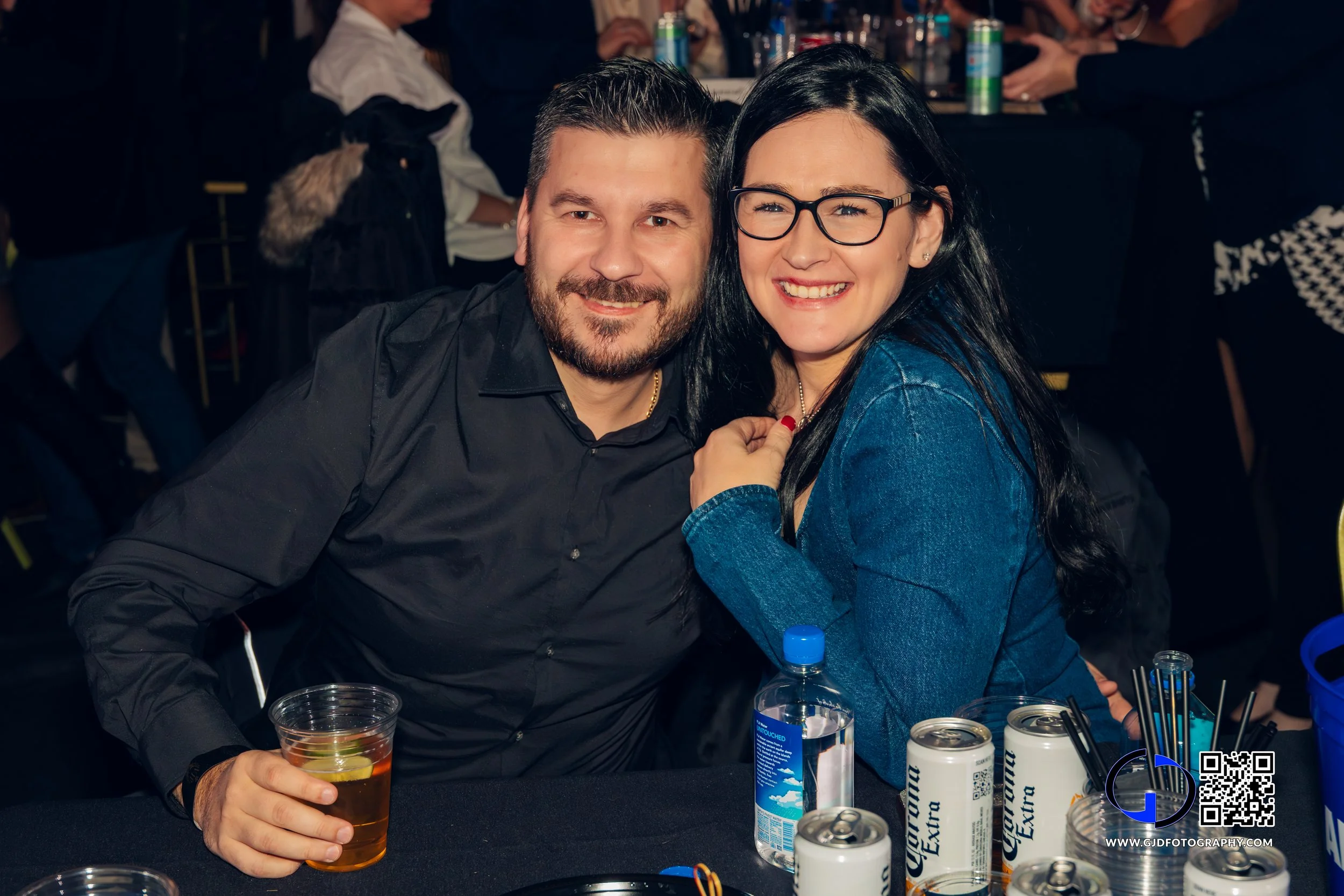 A man and woman smiling at a party seated at a table with drinks, including beer cans, a water bottle, and a glass of whiskey, in a crowded indoor setting.