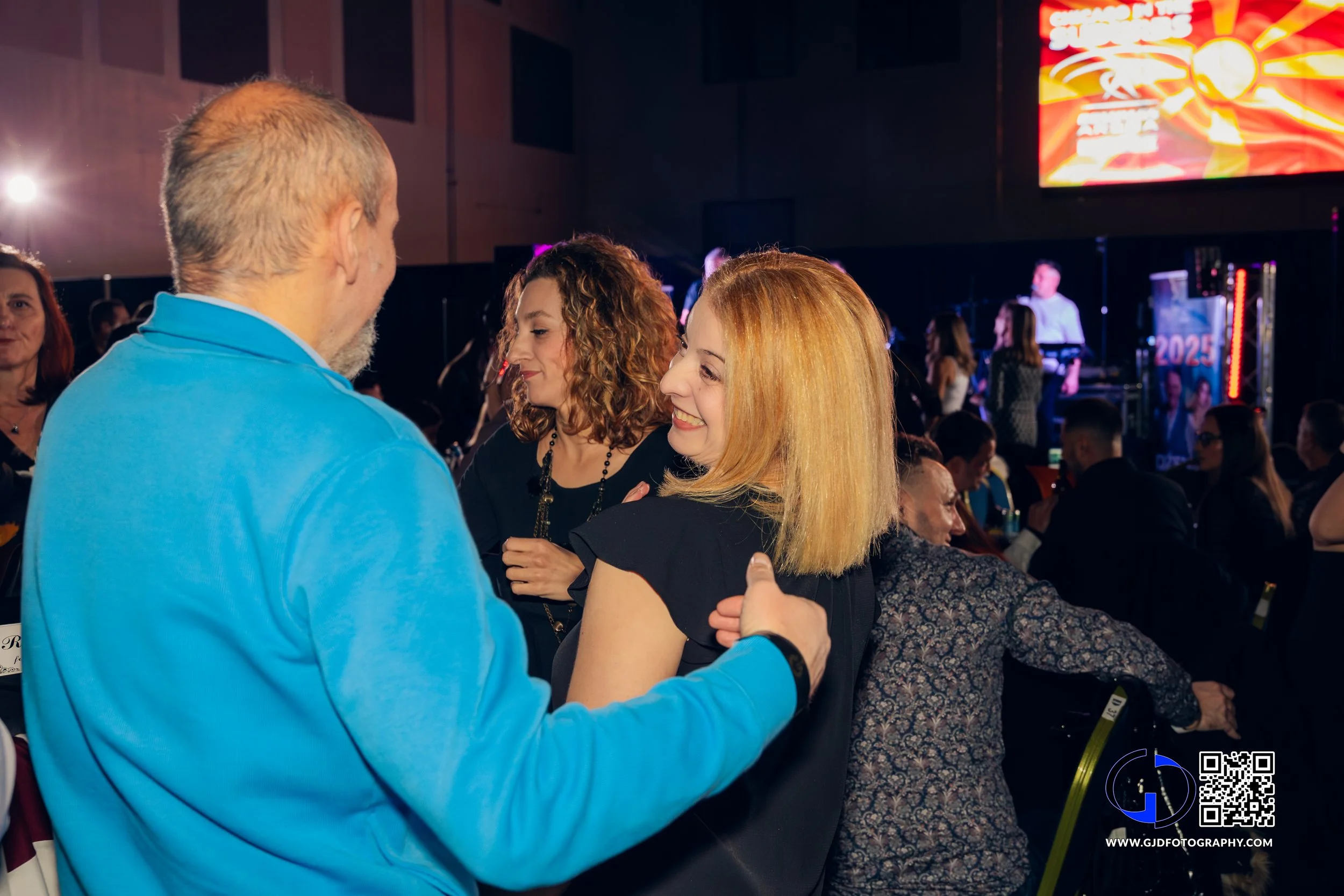 Group of people talking and laughing at an indoor event or concert, with a stage and bright screen in the background.