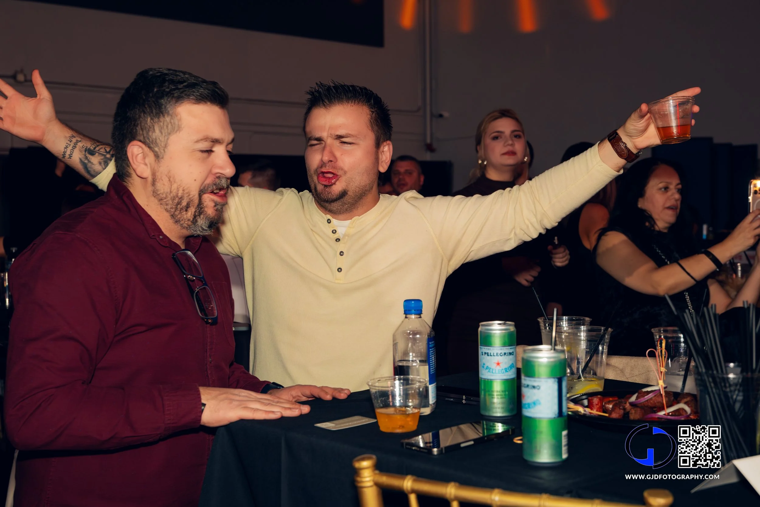Two men are dancing and singing at a party, one wearing a yellow shirt with arms outstretched, the other in a maroon shirt with glasses hanging from his collar. There are drinks on the table.