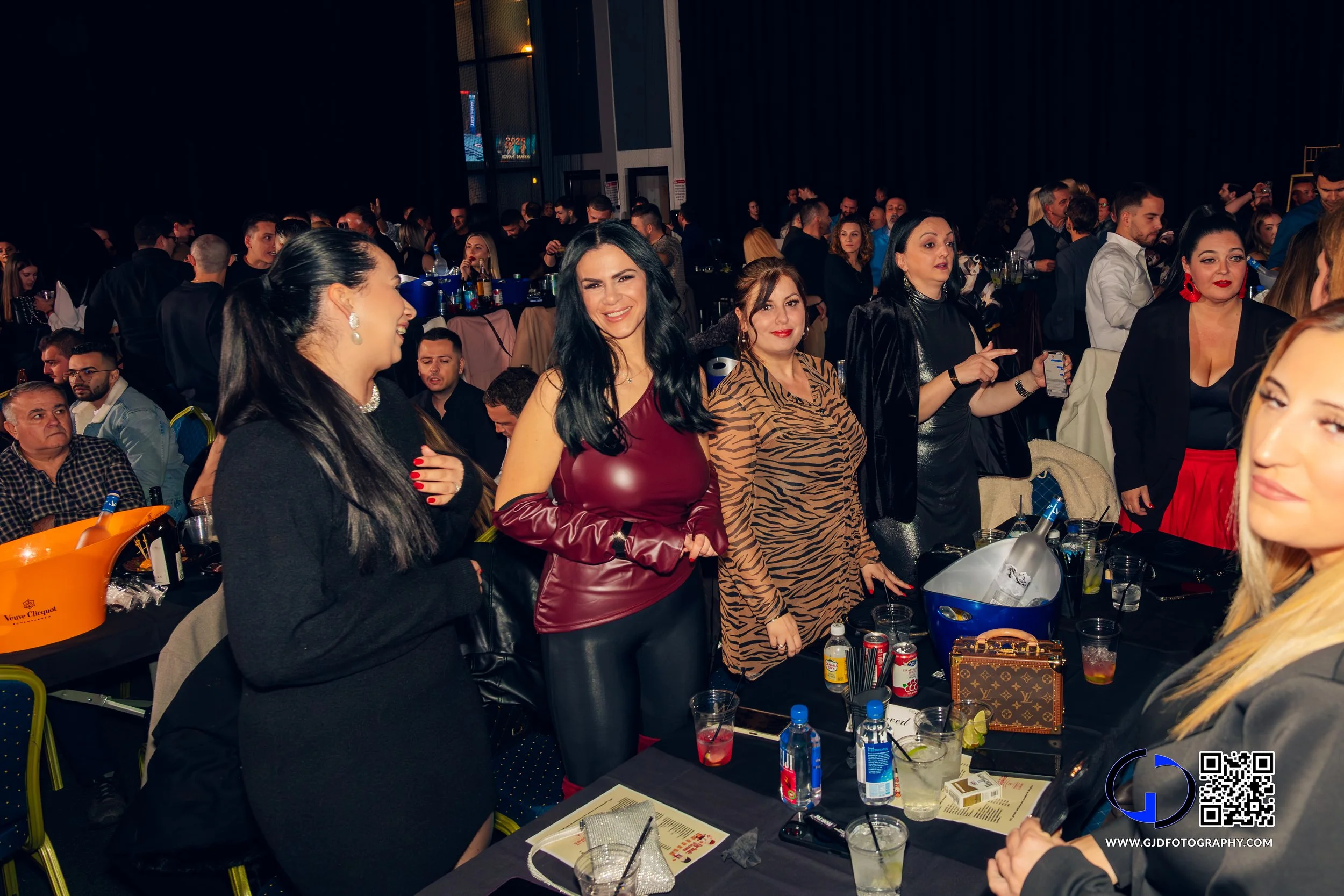 A group of women standing at a table in a large event hall, smiling and engaging with each other, with many people seated and mingling in the background.