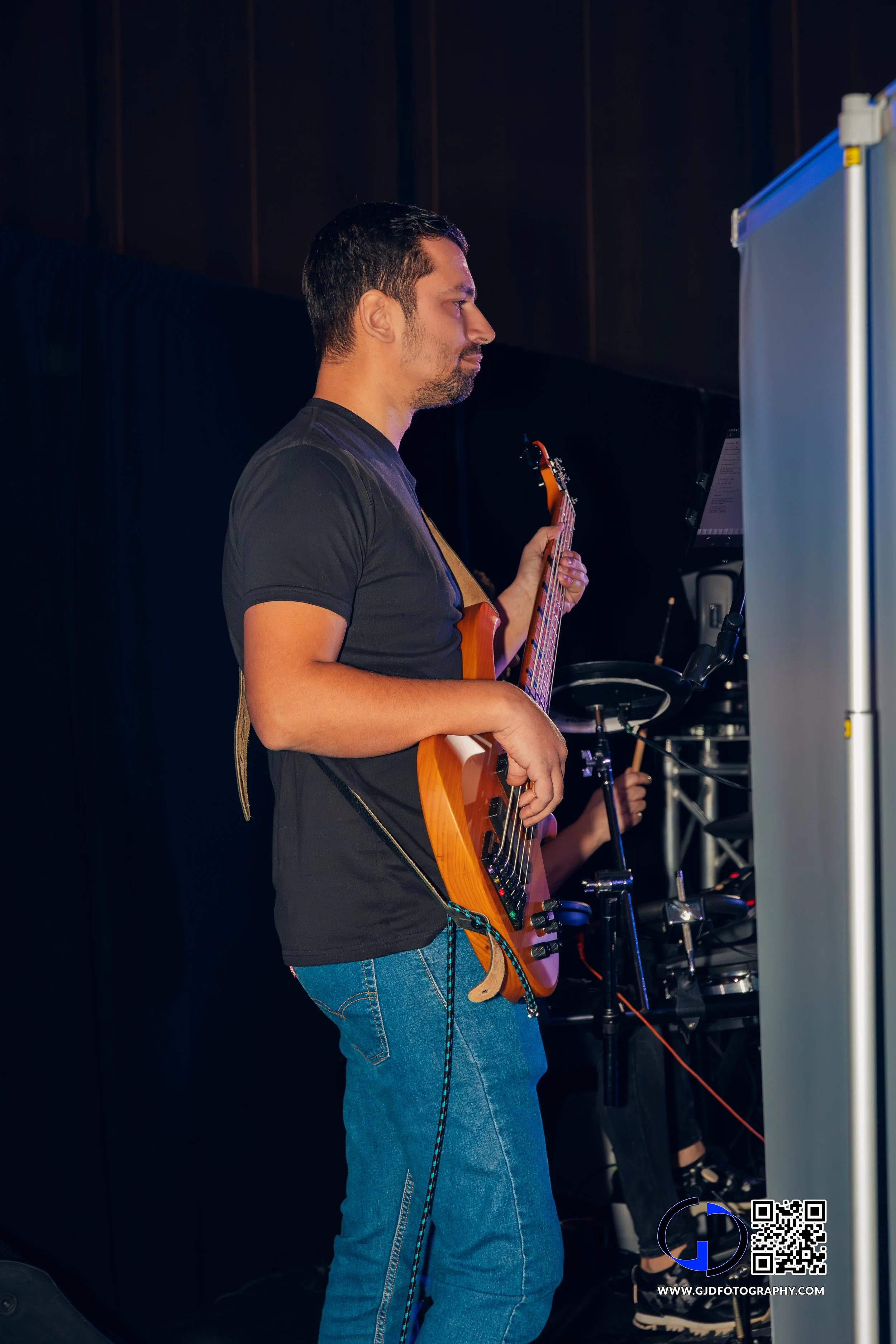 A man playing an electric guitar on stage, wearing a black t-shirt and jeans.