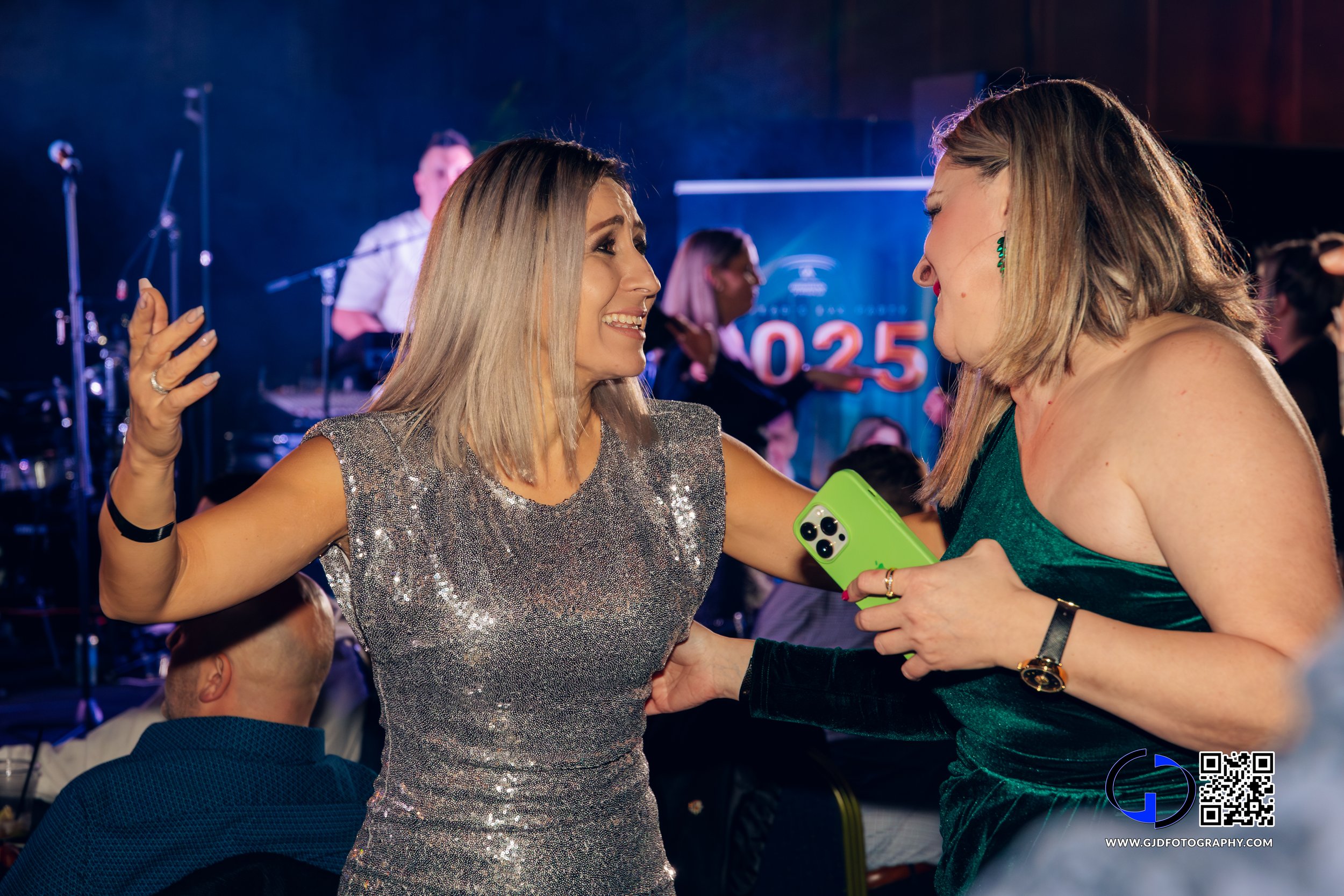 Two women are animatedly talking and smiling at each other at a lively event. One woman is wearing a glittery silver dress and the other is in a green velvet dress, holding a green phone. There are people and a stage with musical instruments in the b