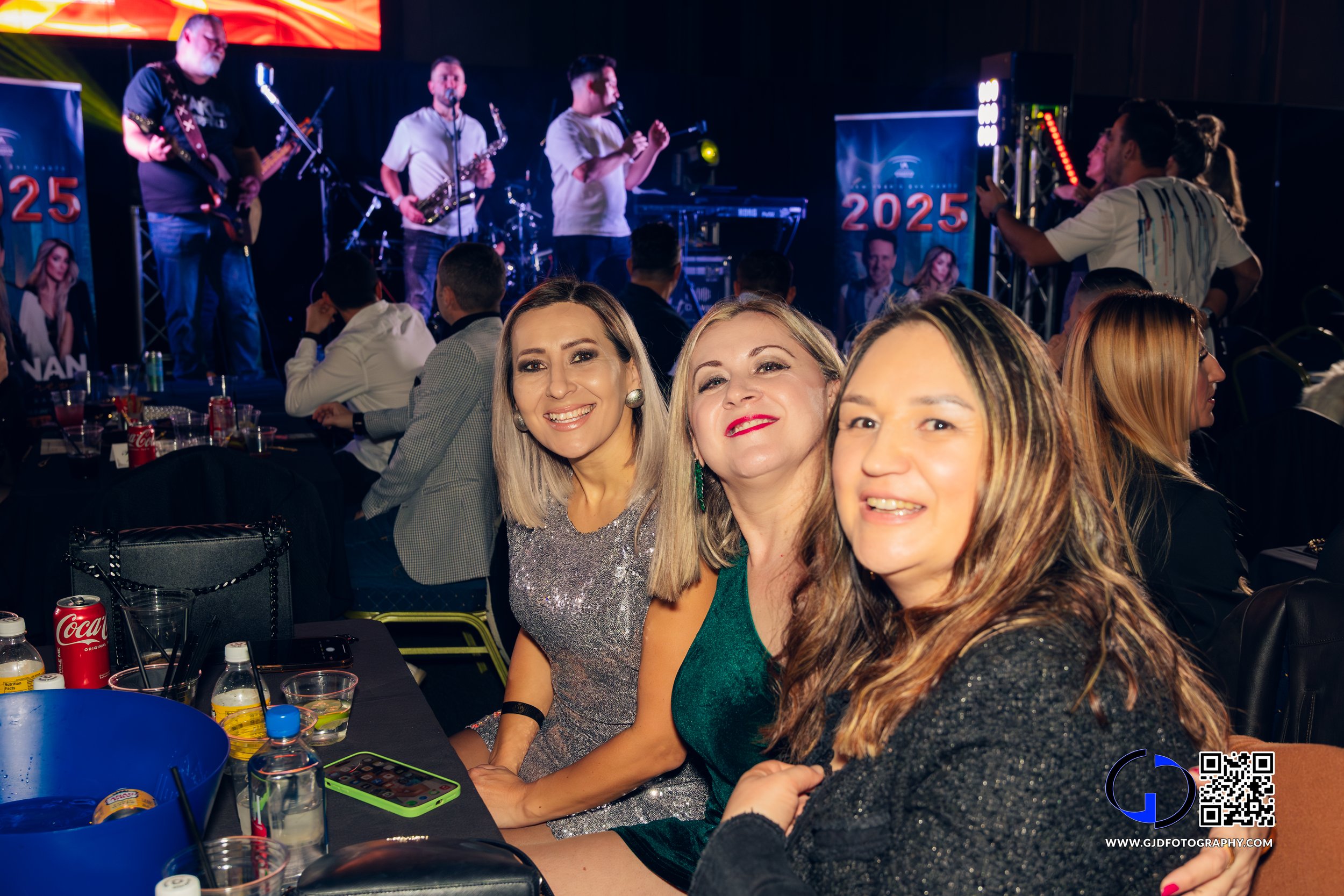 Three women smiling and sitting at a table during a live music event with a band performing on stage in the background.