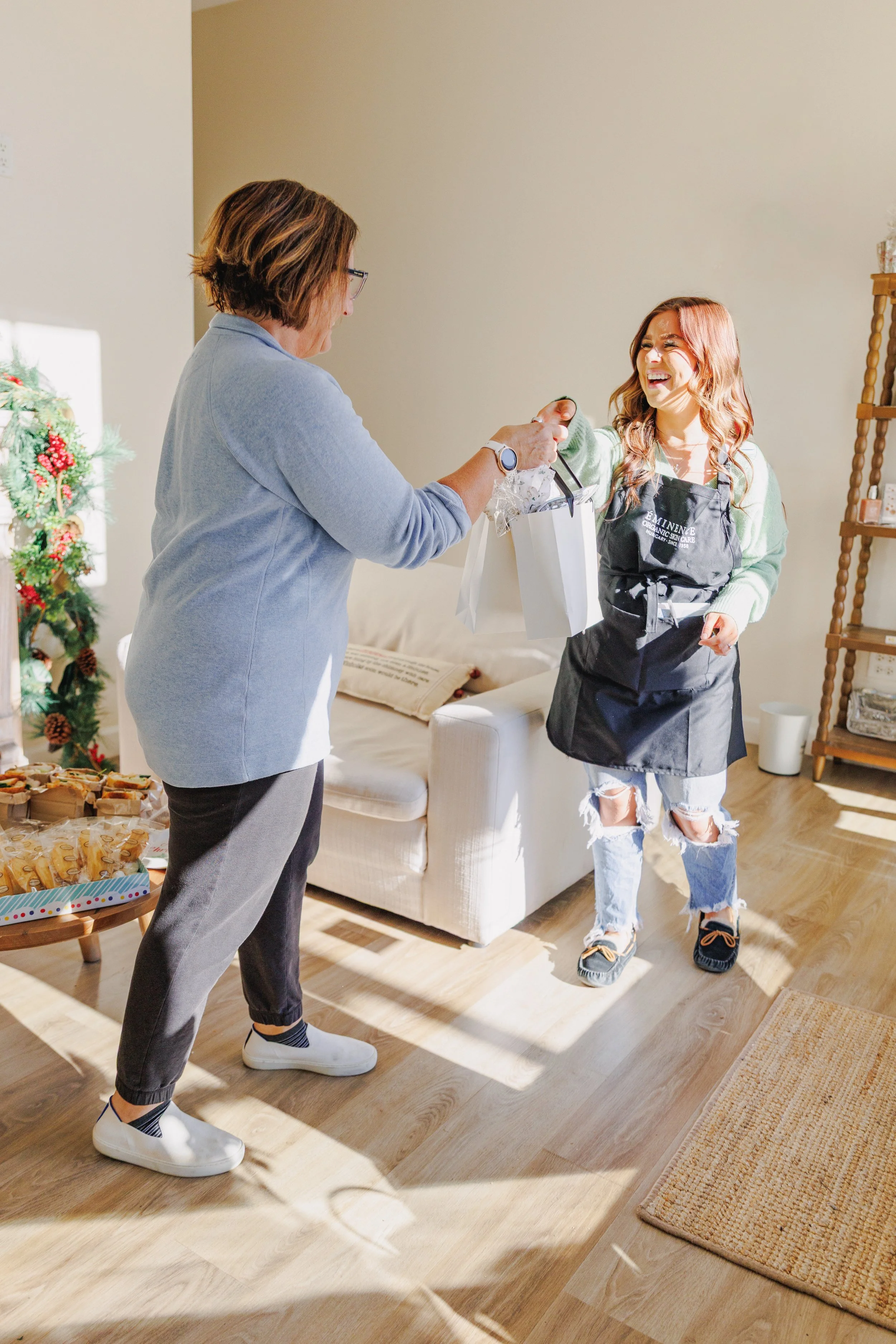 A woman receiving a gift from another woman at a holiday gathering in a living room with a decorated Christmas tree and snacks on a table.