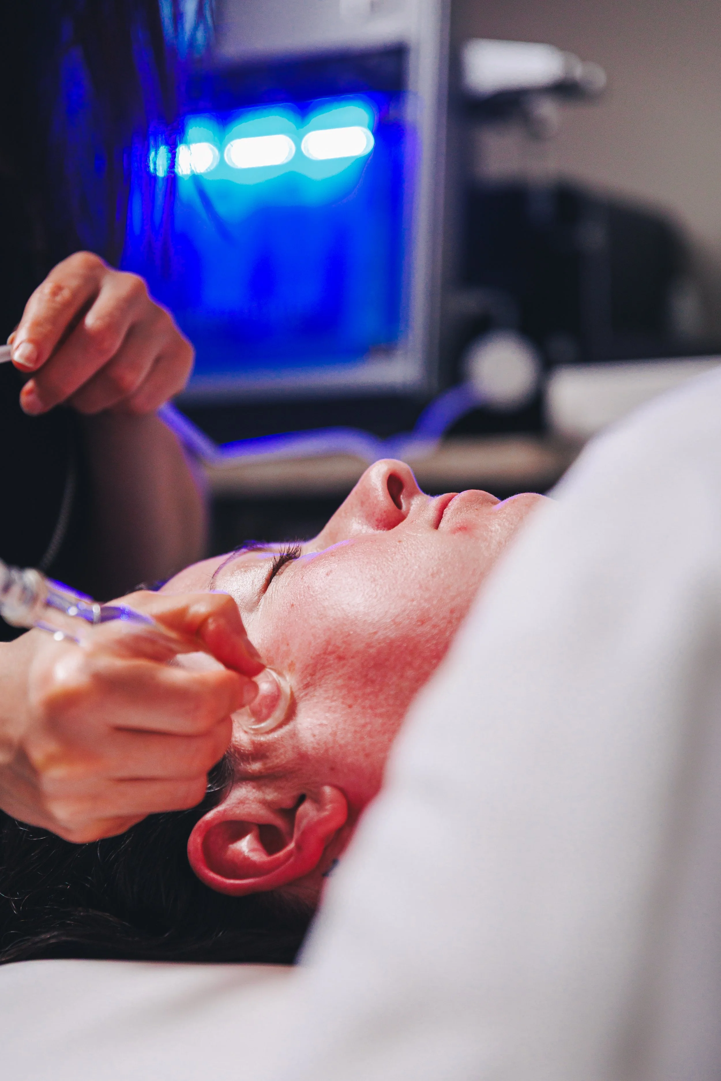A woman receives a cosmetic injection near her eyebrow, with a medical professional holding a syringe, in a clinical setting with a computer monitor displaying blue light in the background.