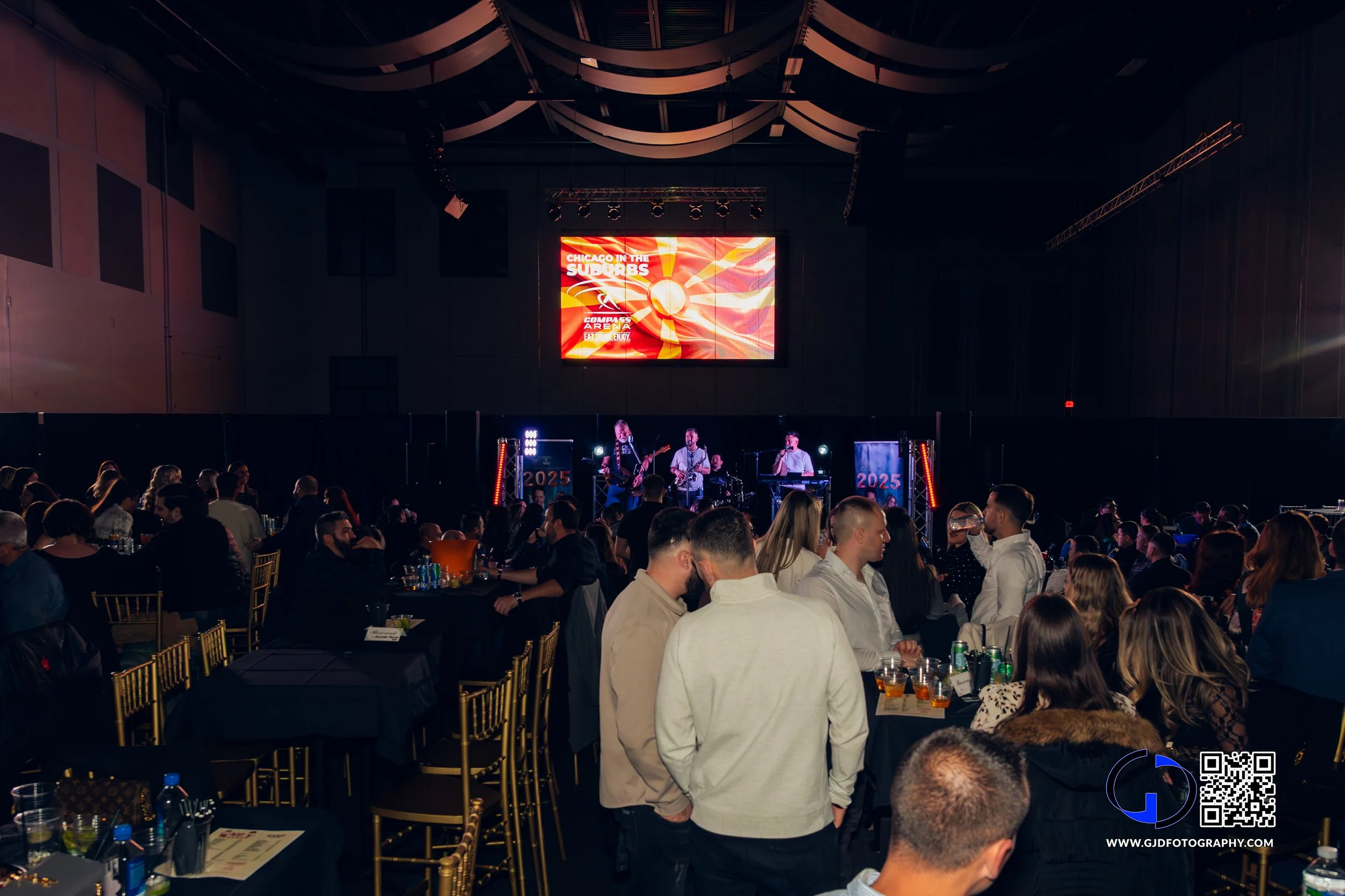 Concert event with a band performing on stage in a large indoor venue, audience seated at tables, with a large screen displaying 'Chicago in the Suburbs' and 'Compuware Arena' in the background.