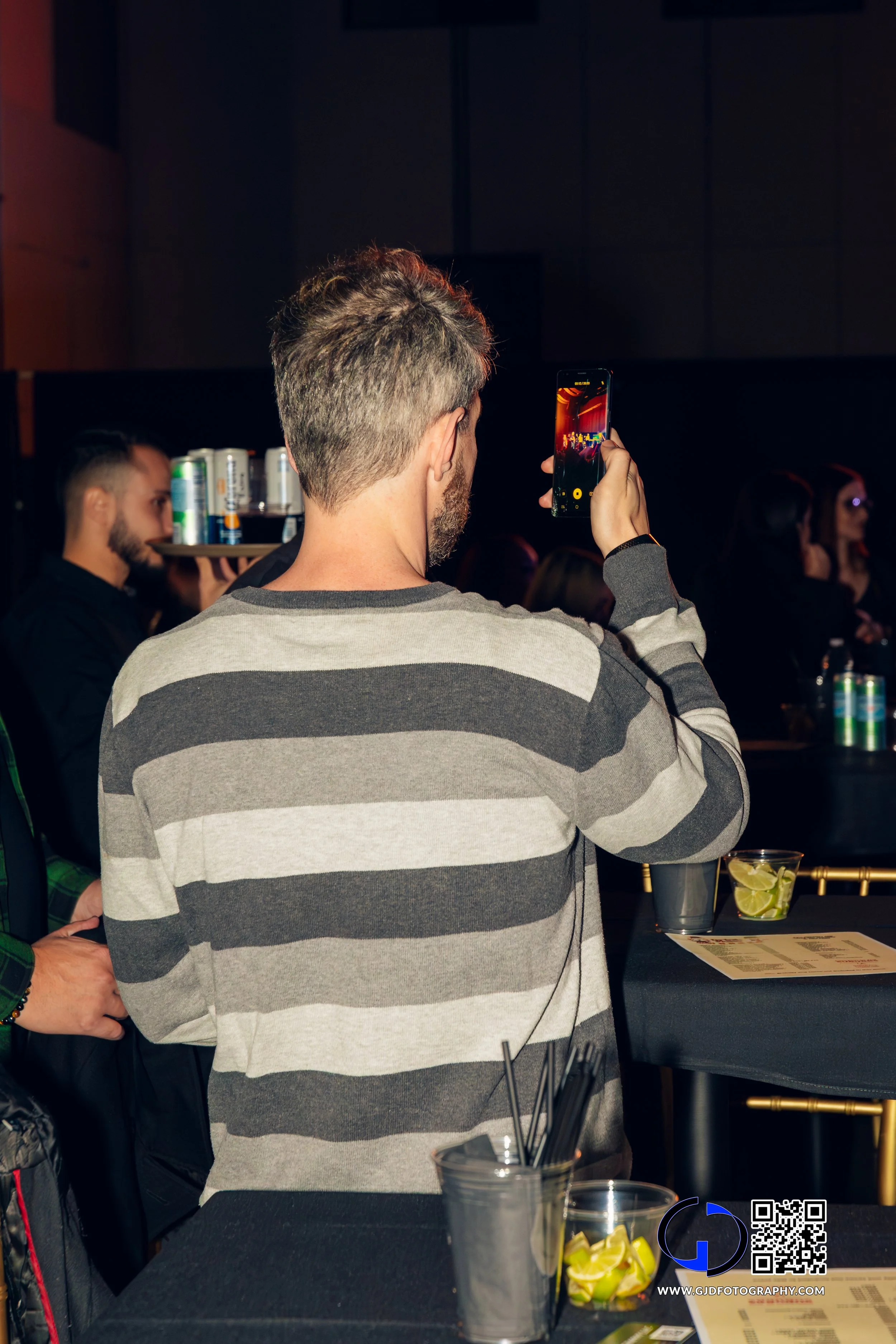 A man with short hair and a beard wearing a gray and black striped shirt takes a photo with his smartphone at a social event. There are drinks, lime slices, and a menu on the table, with other people in the background.