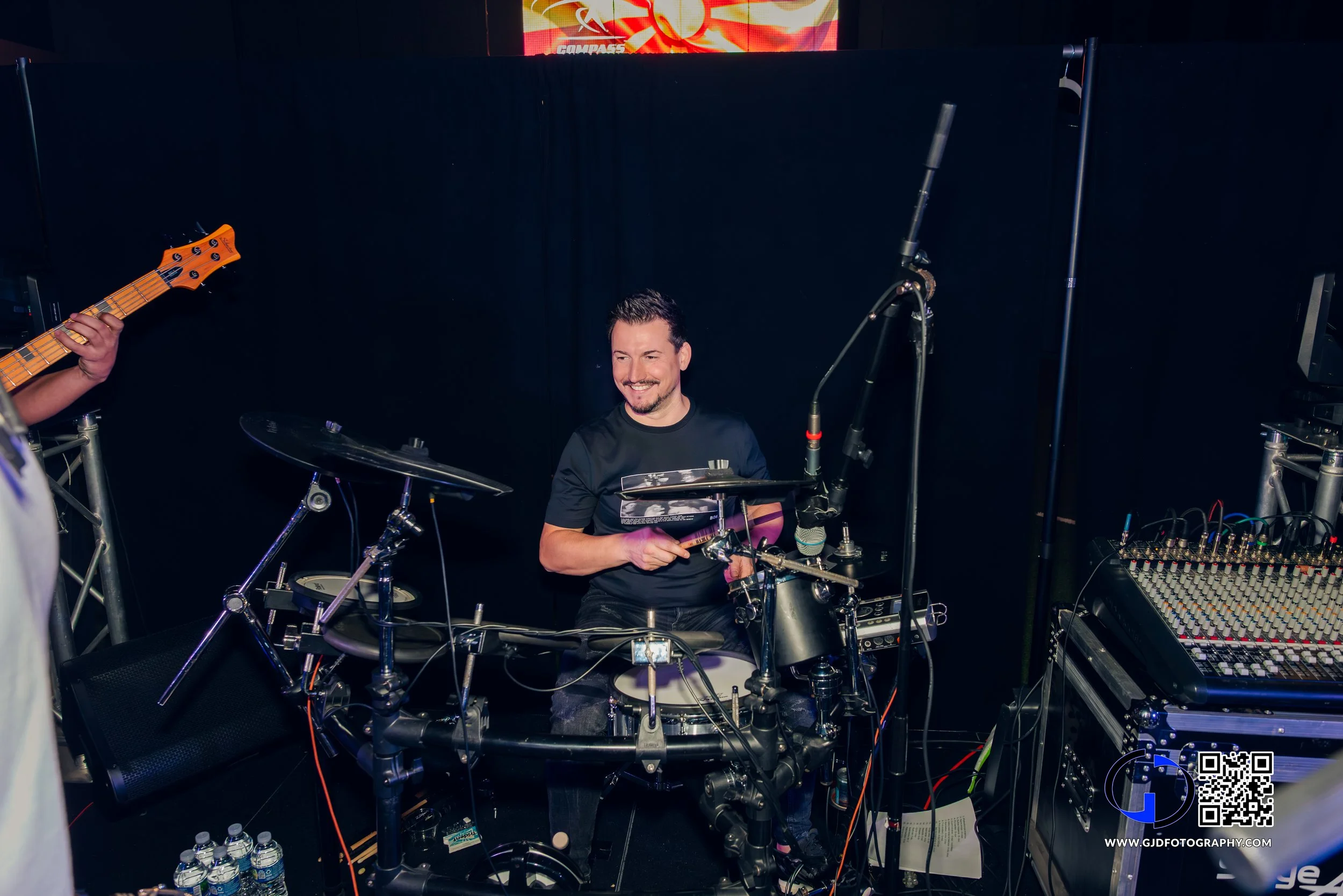 A man smiling and playing an electronic drum set on stage with a sound mixer to his right and a person playing an electric guitar to his left. There is a black backdrop and a digital display above with some graphics.