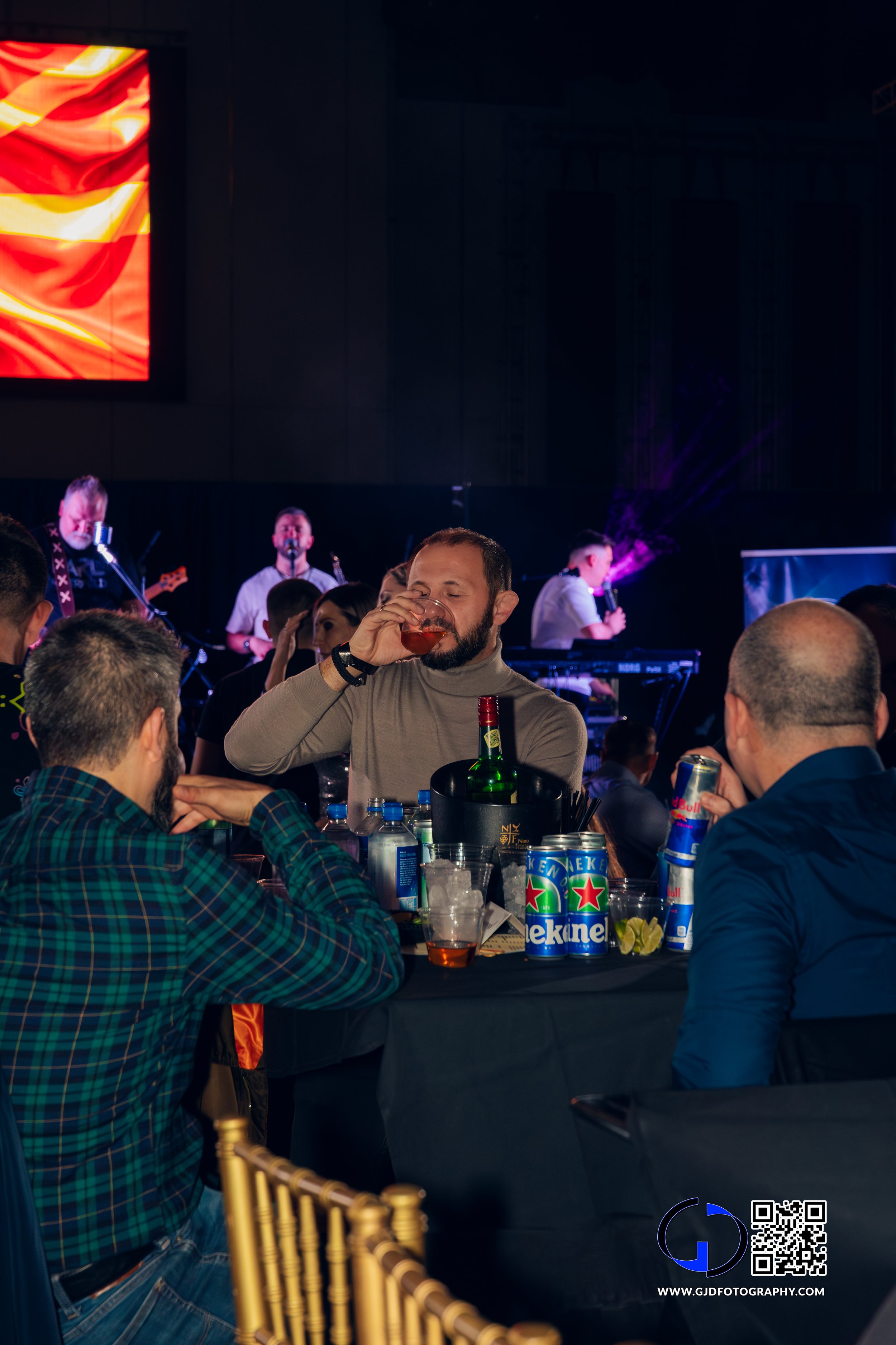 A man with a beard and gray turtleneck drinks from a glass at a table surrounded by other people at a lively indoor event with a band performing on stage in the background, and drinks including Heineken beer, Red Bull energy drinks, and water bottles