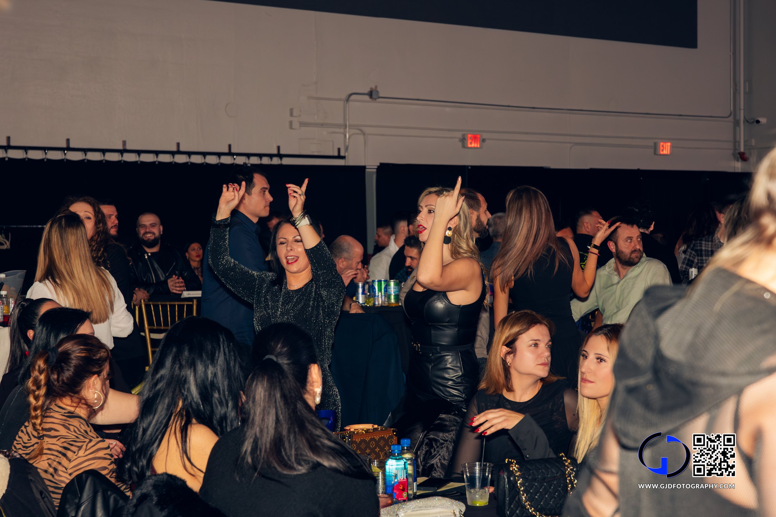 People dancing and socializing at a party or event, with some sitting and some standing, in a dimly lit room with black walls.