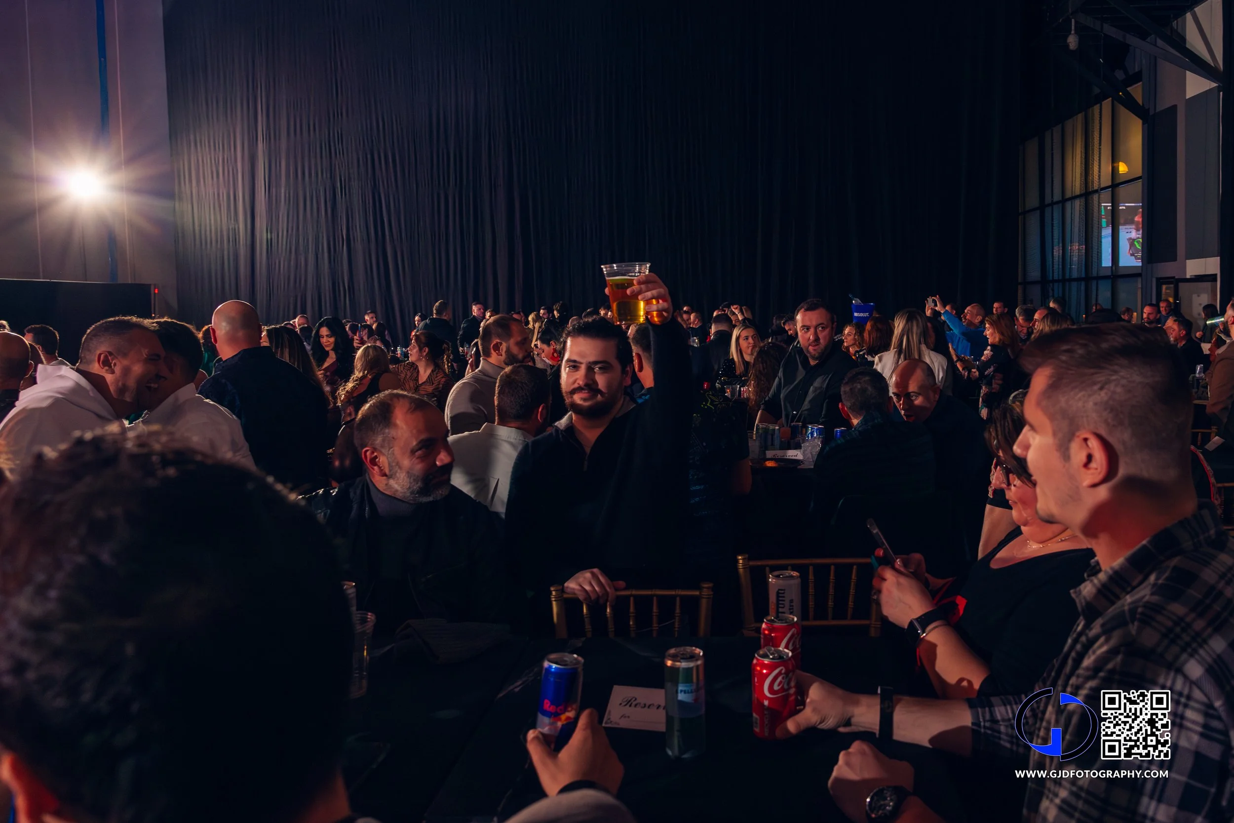 Crowd of people at a lively event or party, some seated at tables, one man in the center holding up a glass of beer, dim lighting with a dark backdrop, and some people using their phones.