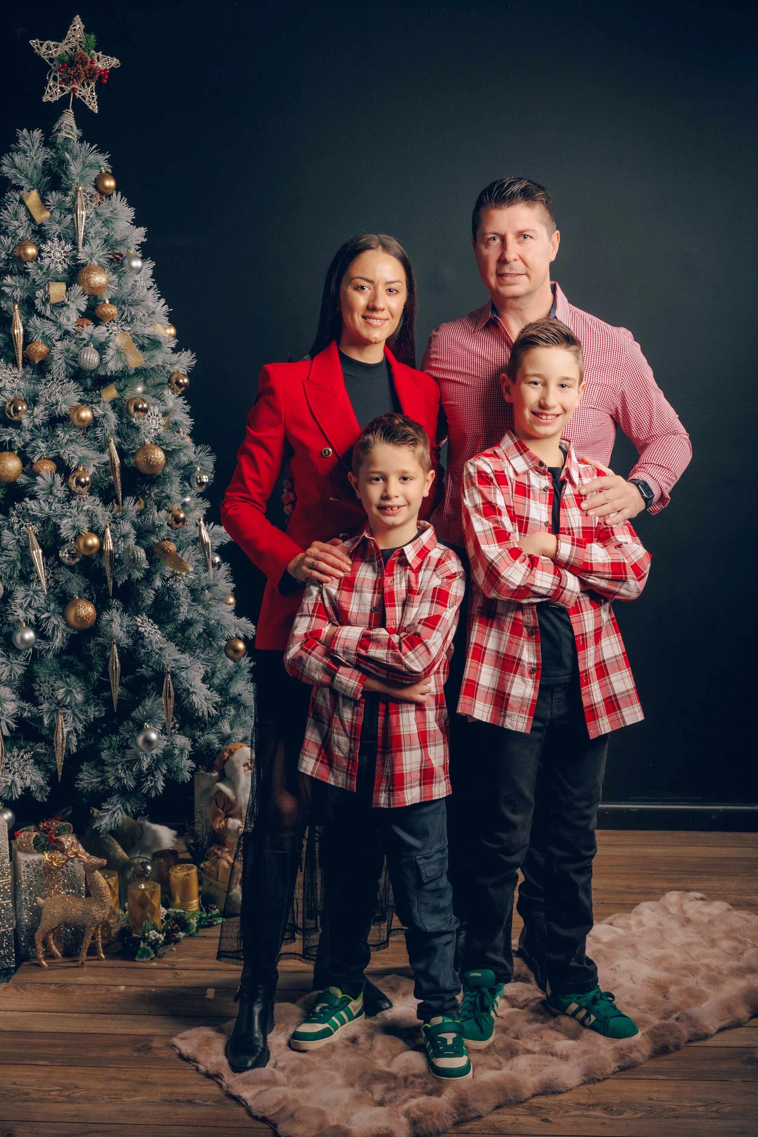 A family of four standing indoors near a decorated Christmas tree, with a dark wall in the background and a furry rug on the wooden floor.