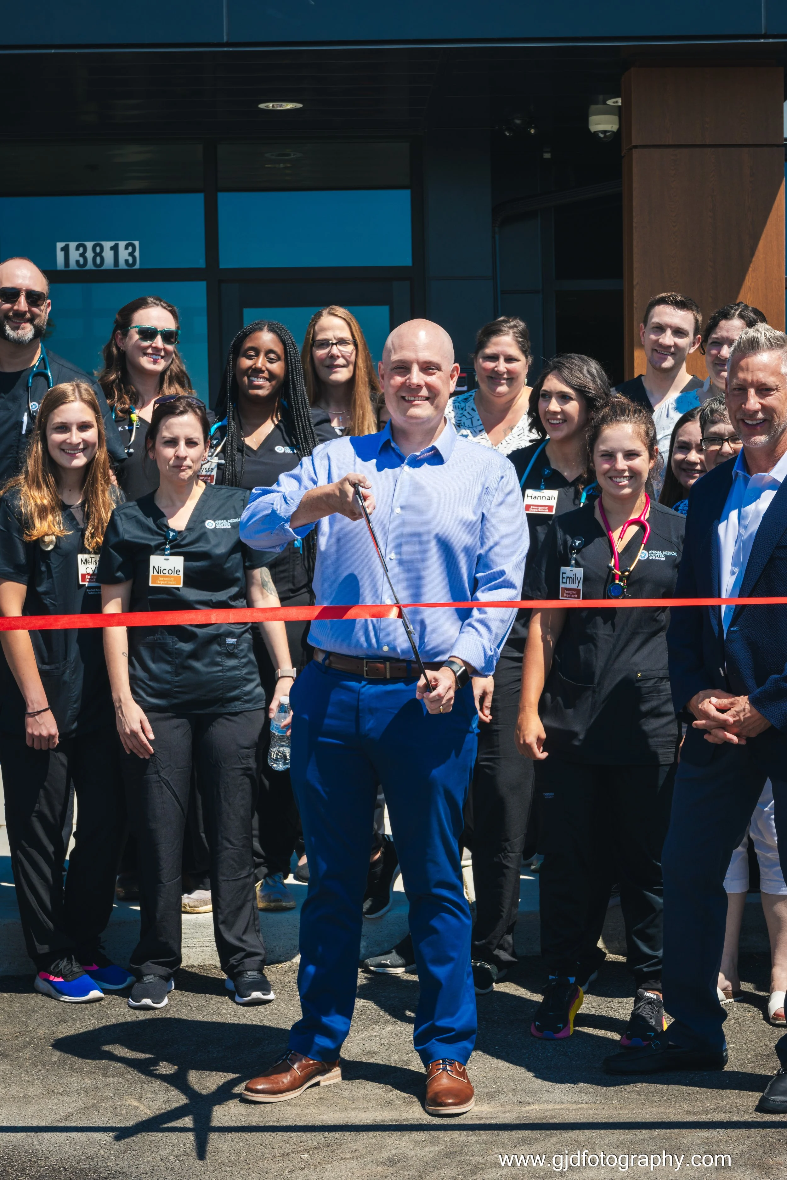 A man in a blue shirt and blue pants cuts a red ribbon in front of a group of people at a ribbon-cutting ceremony outside a building with the number 13813. The group includes healthcare workers and officials smiling for the photo.