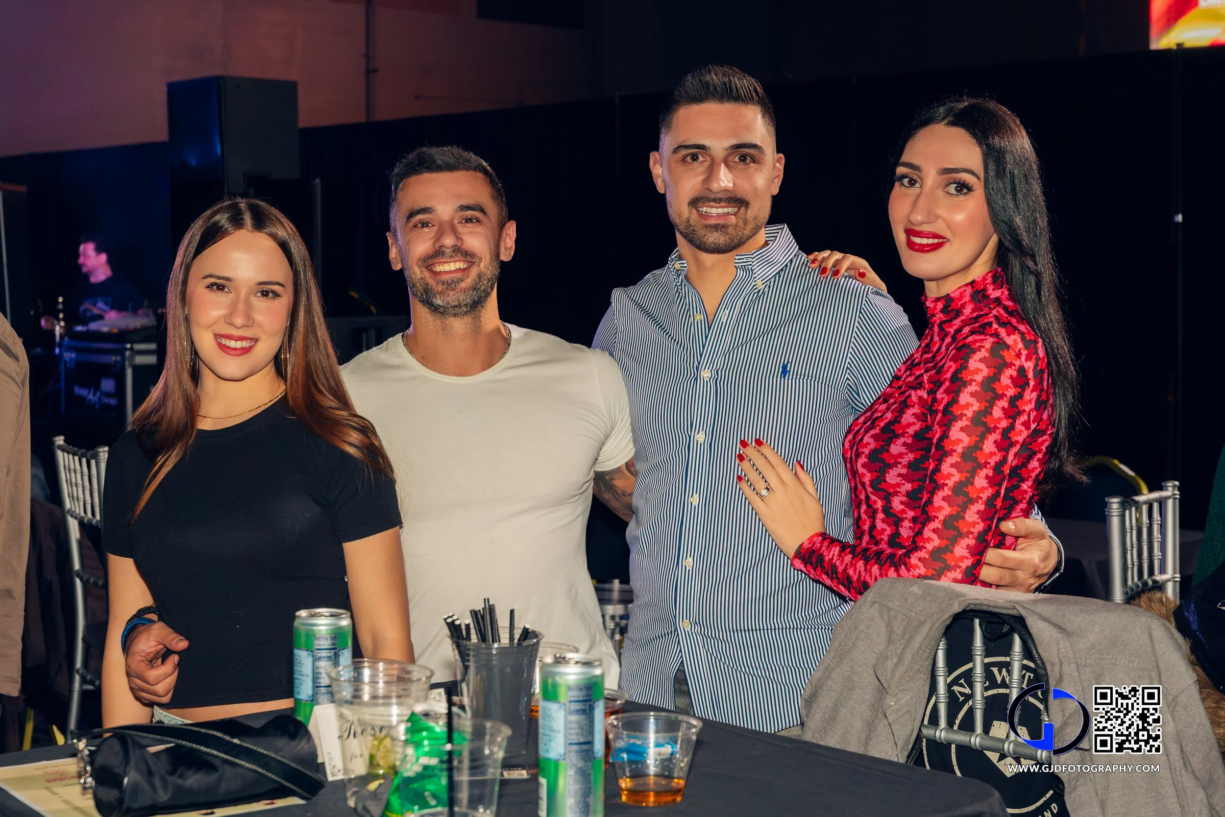 Four friends smiling, standing close together at a table during a social event or party.