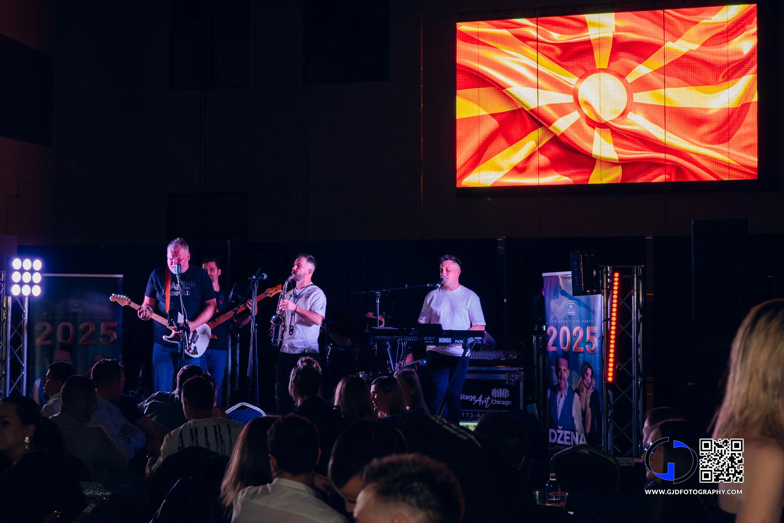 A band performing on stage with a digital screen displaying a graphic of the North Macedonia flag, an audience watching, and banners for 2025 event with people dressed in formal and casual attire.