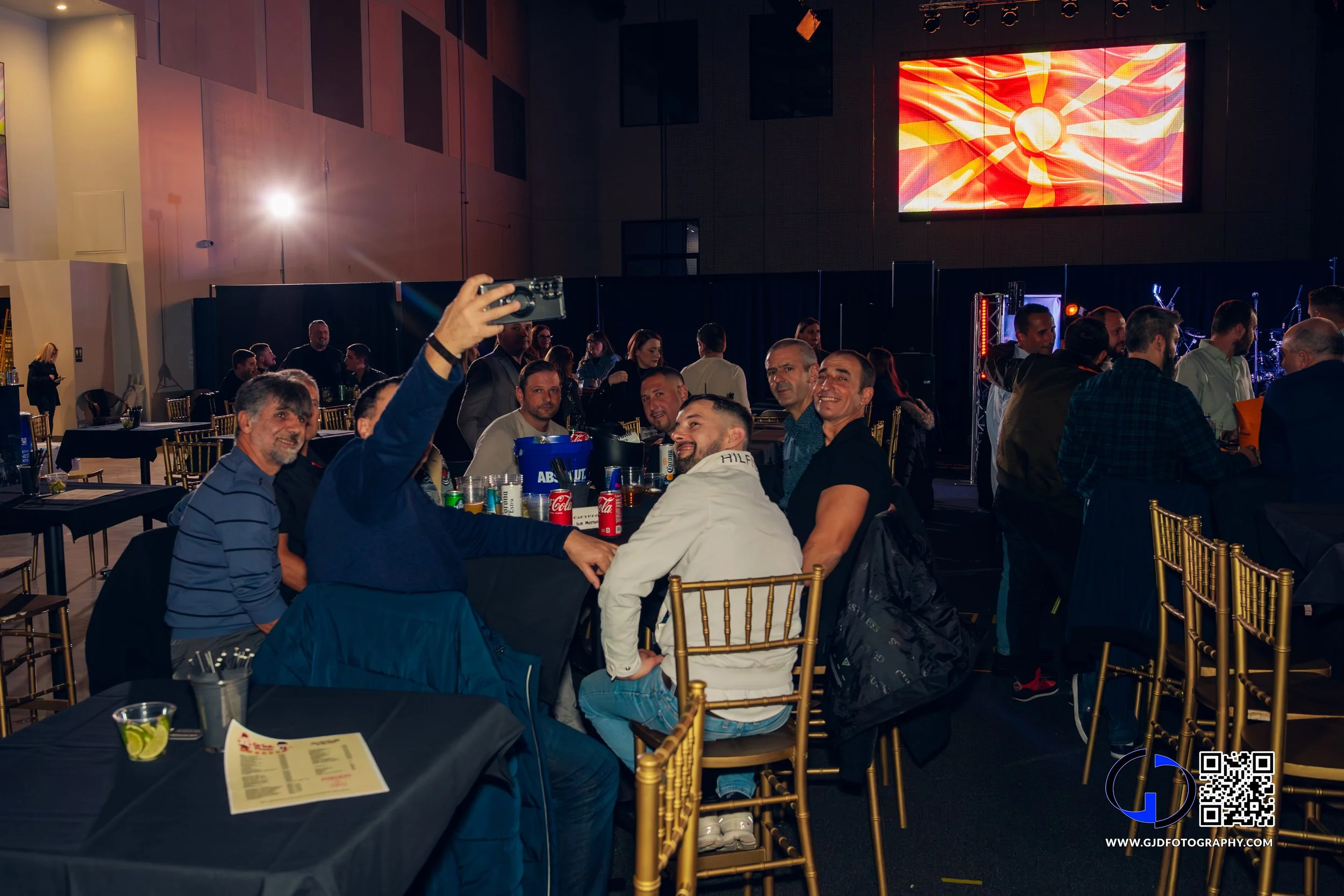Group of people sitting at a table in a banquet hall taking a selfie, with a large screen displaying a bright sun-like graphic in the background.
