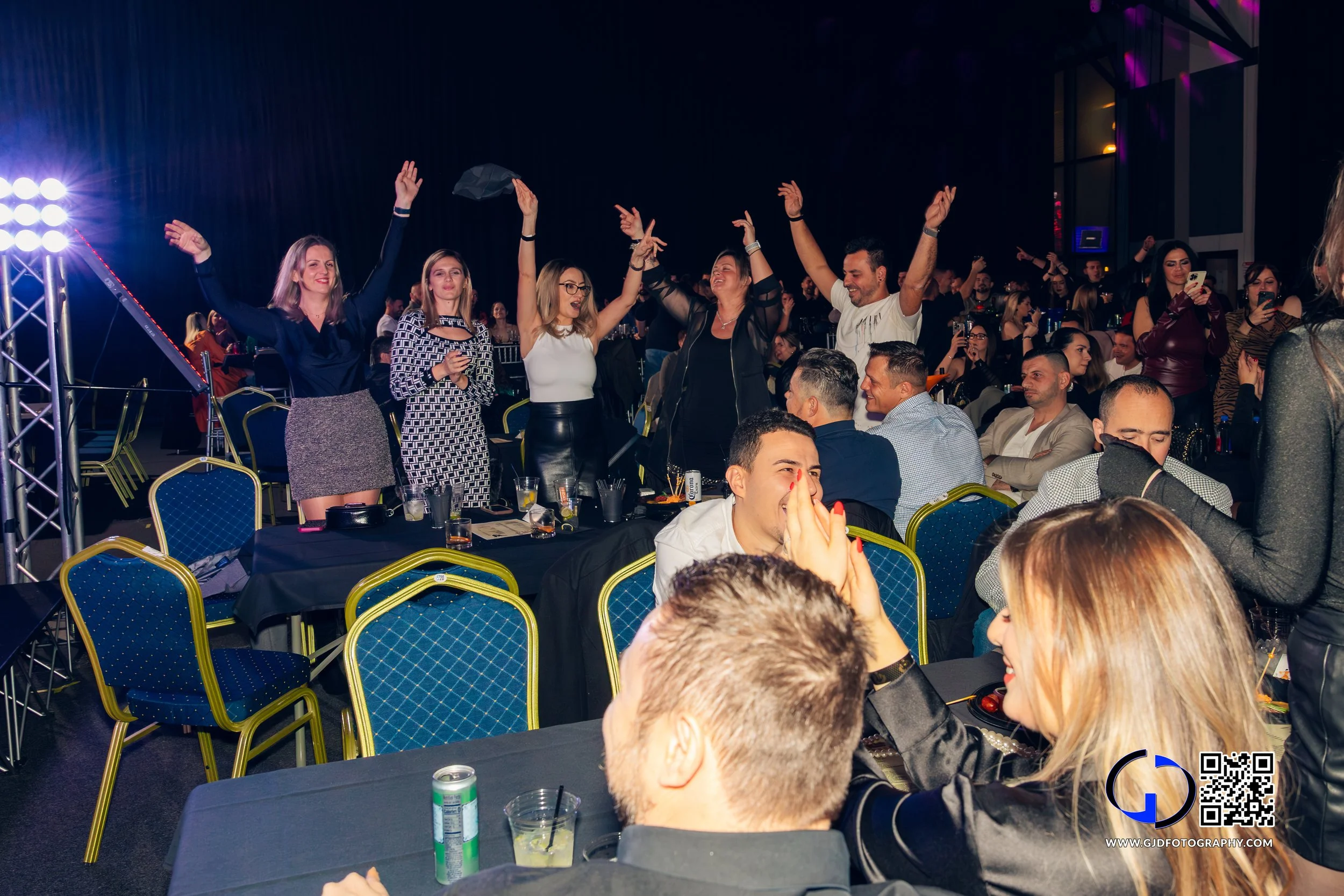 People dancing and socializing at a lively indoor event with tables, drinks, and a stage with bright lights in the background.