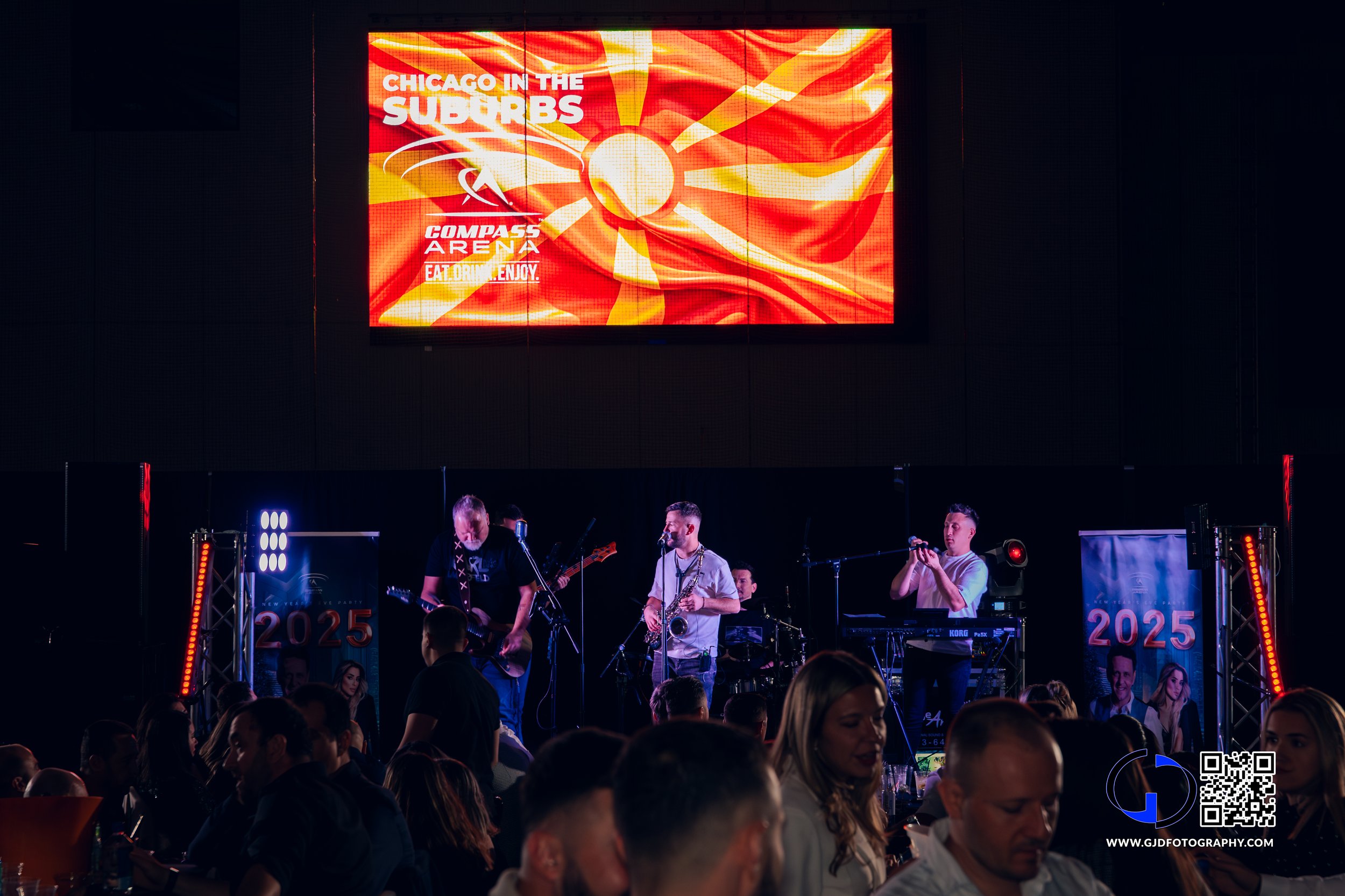 A band performing live on stage under a large LED screen showing a graphic that says "Chicago in the Suburbs" and "Compass Arena" with a bright, abstract sun design. The audience is seated in front of the stage.