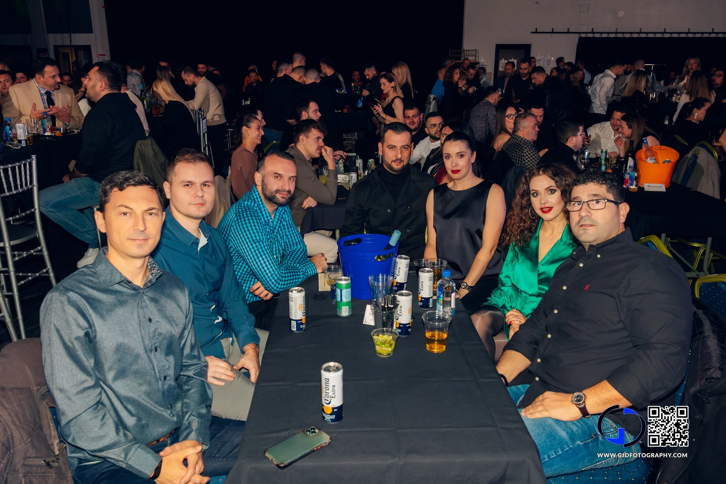 Group of eight people seated around a table at a large indoor event, with many other people in the background, drinks on the table, and a black tablecloth.