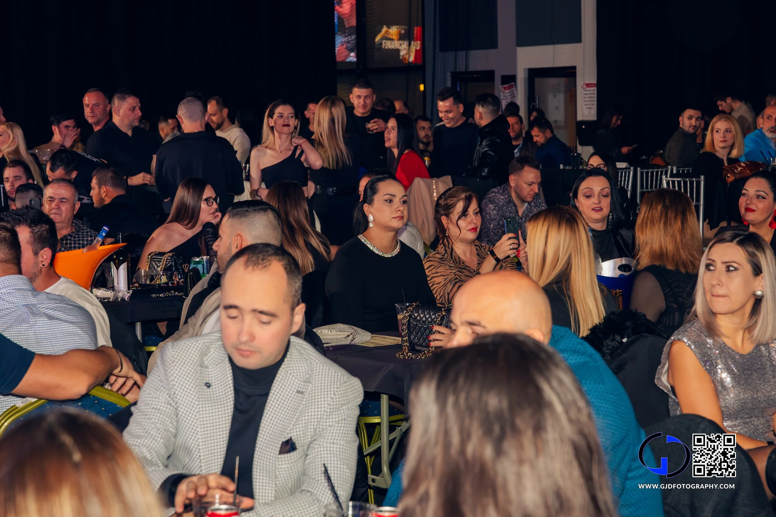 Crowd of people sitting and standing at an indoor event, some using their phones, with a woman in a black dress with pearl necklace in the center.