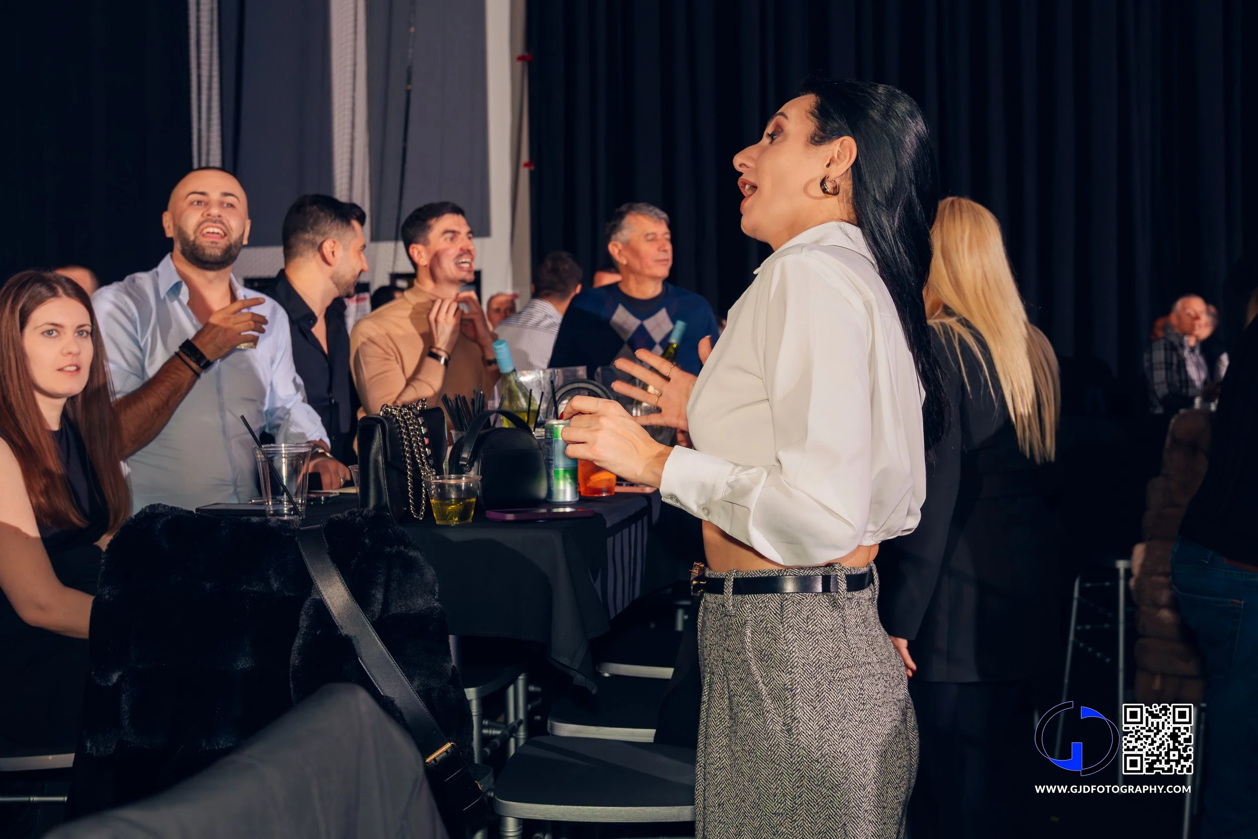 A woman in a white blouse and gray plaid pants talking to a group of people at a bar or lounge, with drinks on the table, under dim lighting.