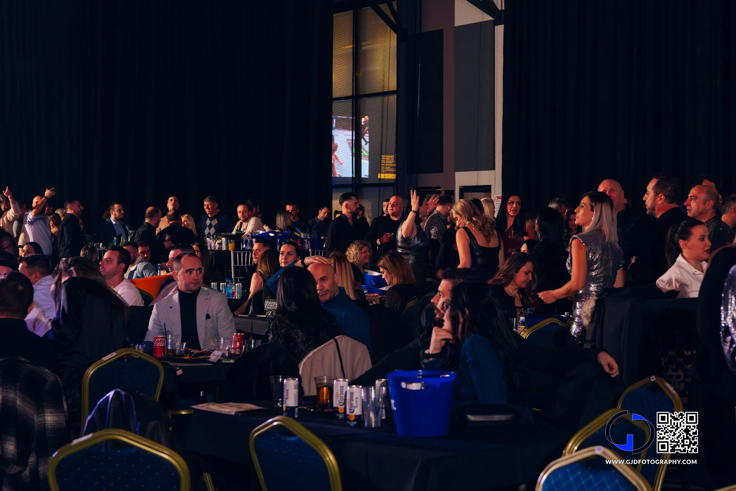 People gather in a dark indoor venue, sitting at tables, socializing and dancing. Some are standing and talking, with a large screen or projection on a wall in the background.