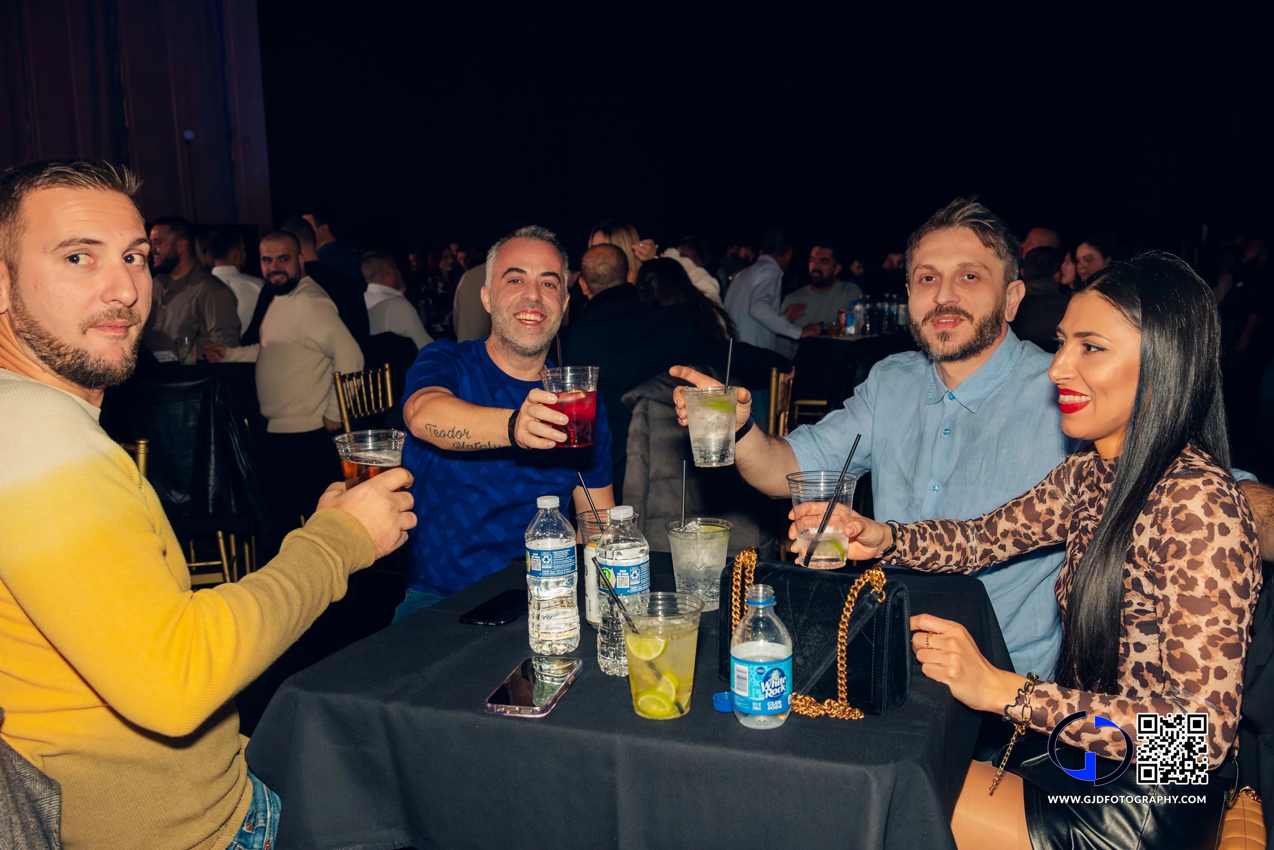 Four friends sitting at a black table in a crowded event, holding drinks and smiling at the camera, with other people in the background.