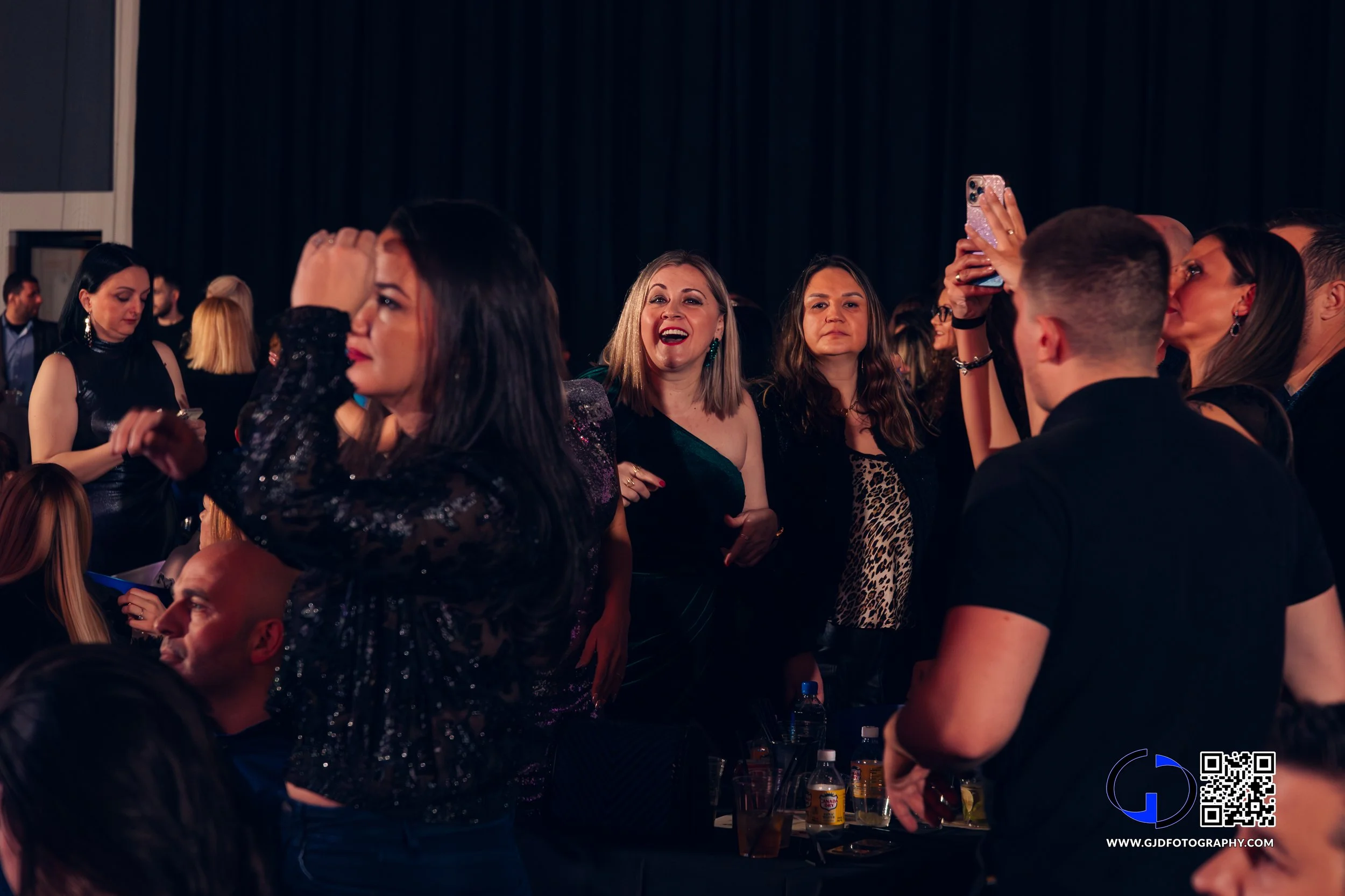 People at a lively party, some dancing and others taking photos, in a dark venue with black curtains.