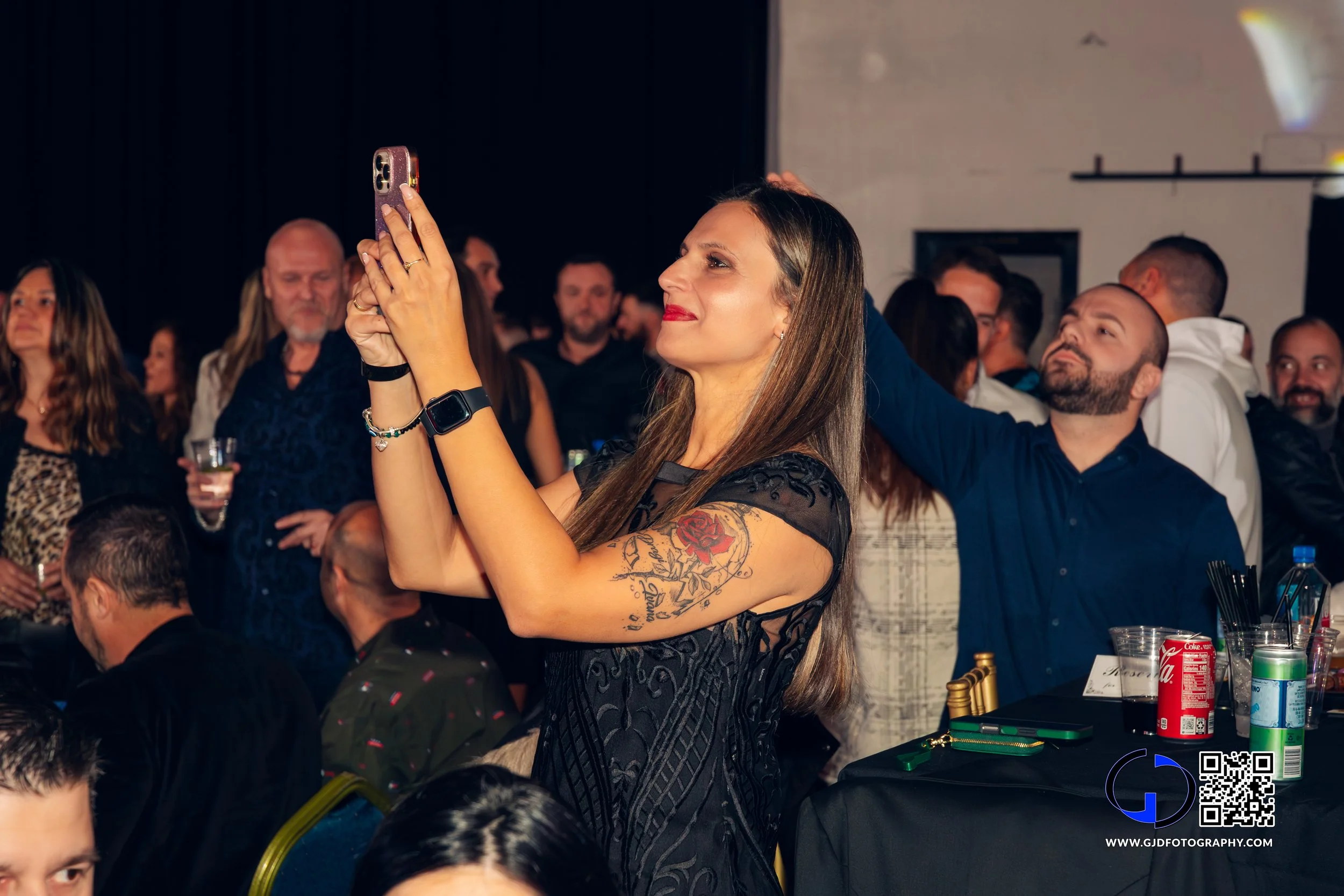 A woman with long hair and a tattoo on her arm holding a phone and taking a photo at a crowded indoor event.