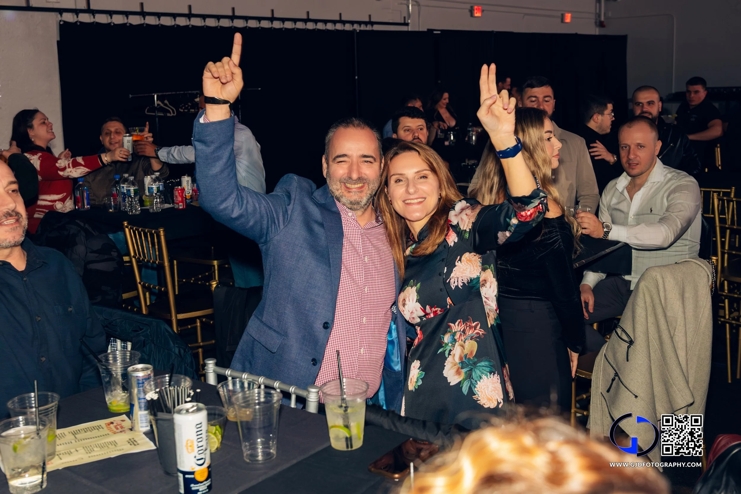 A group of people at a celebration or event, with two people in the foreground smiling and posing with their hands raised. The woman is wearing a floral dress, and the man is in a blue blazer. There are tables with drinks and people enjoying themselv