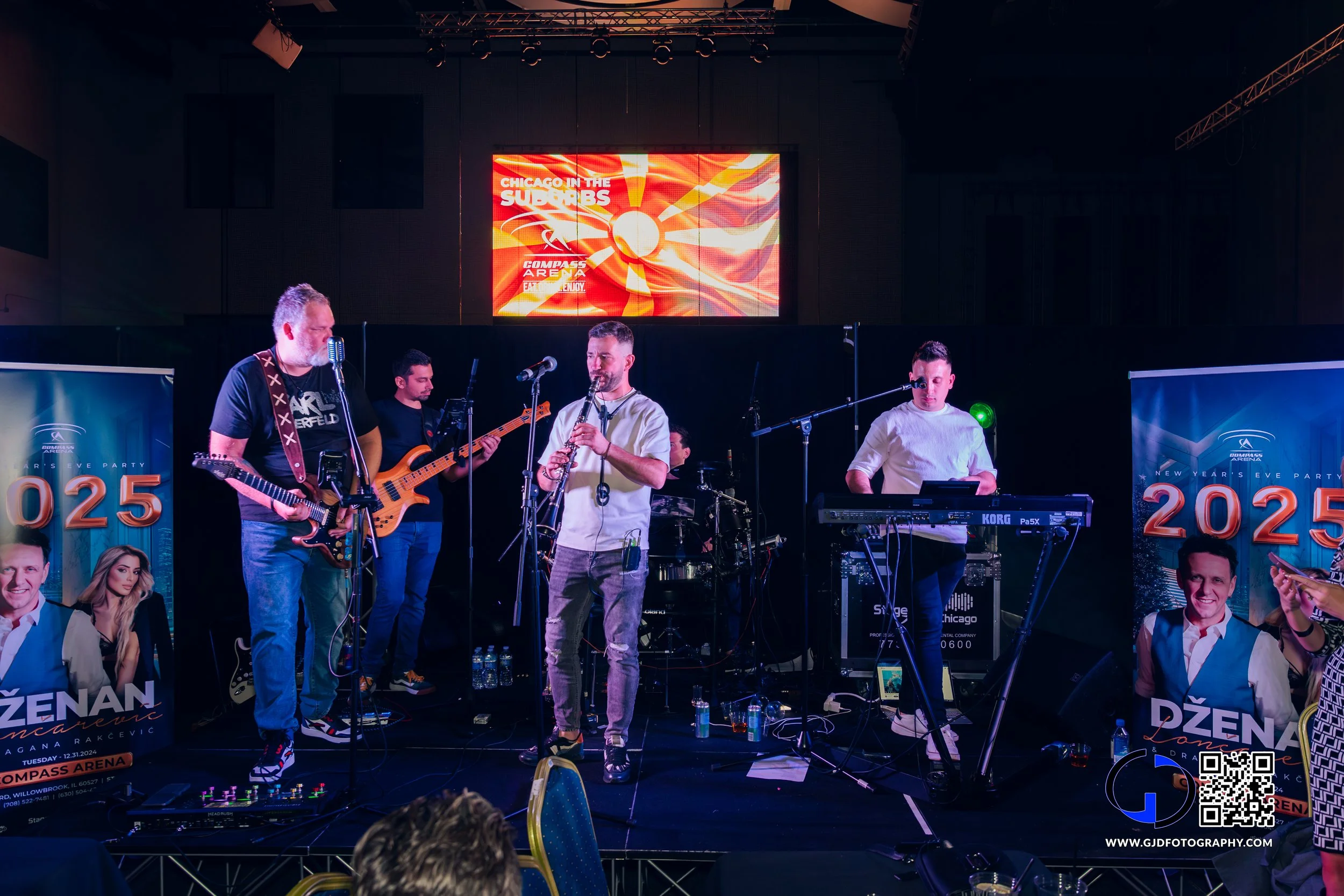 Band performing live on stage at a concert with a large digital screen behind them displaying bright red and orange graphics. The stage has music equipment and promotional banners for a New Year's Eve event at Compass Arena.