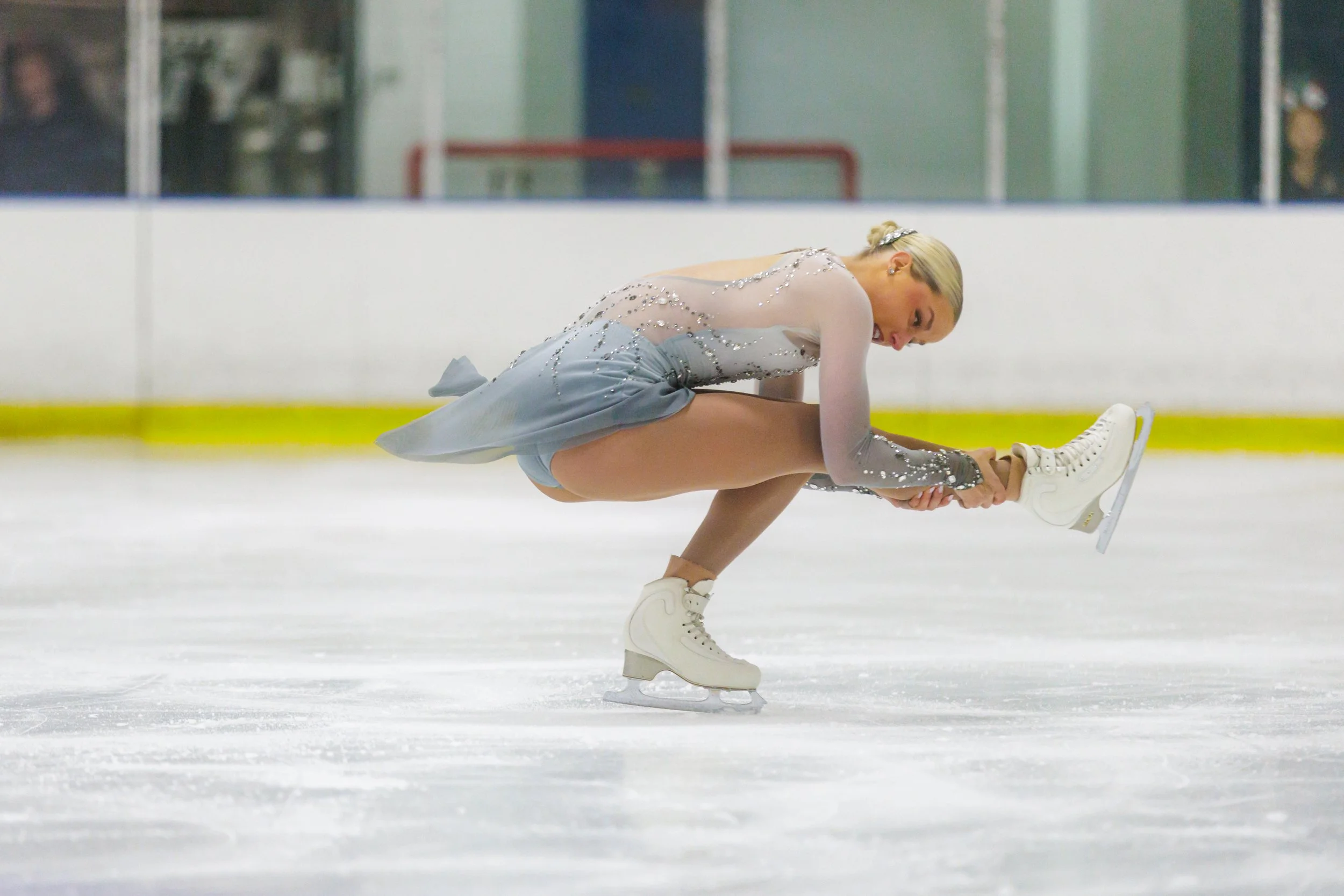 A female figure skater wearing a gray dress with sparkling embellishments performs a move on the ice rink, bending forward while holding her skate blade.